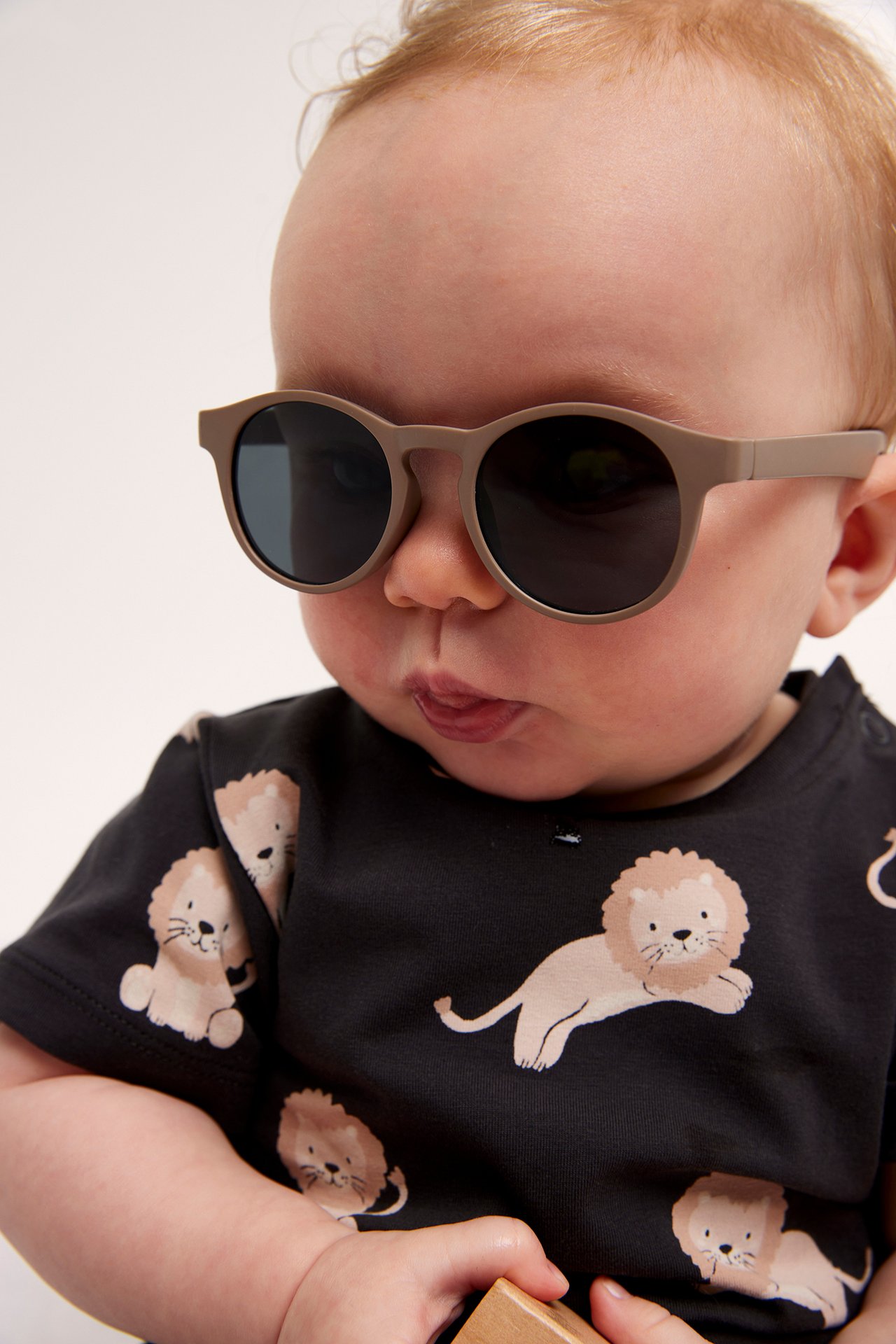 Närbild på en baby som bär beiga solglasögon i plast och en svart t-shirt med lejonmönster.