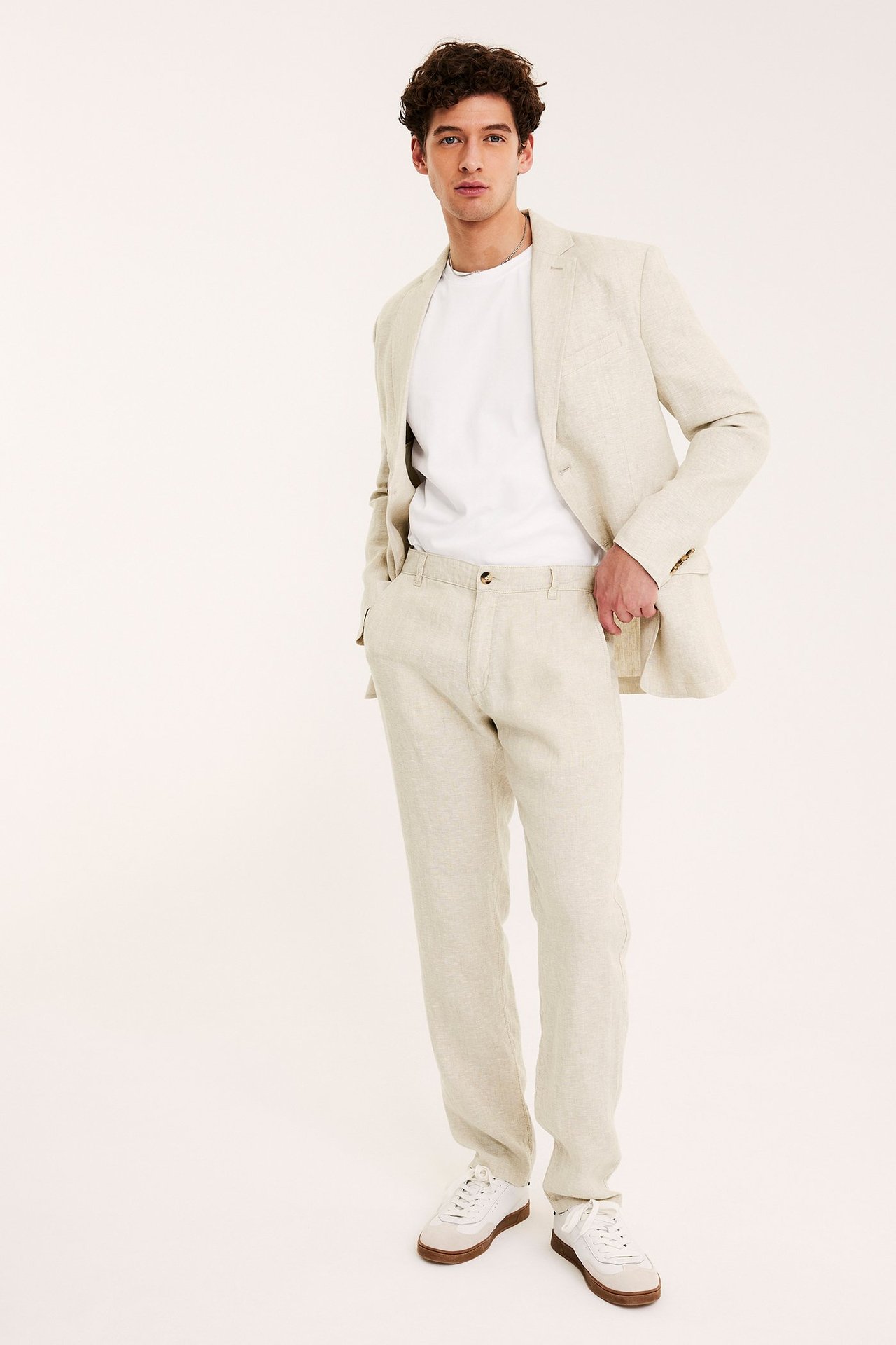 Edestä: mies vaaleanbeiget pellavahousut ja bleiseri yllään, valkoinen t-paita, valkoiset lenkkarit.