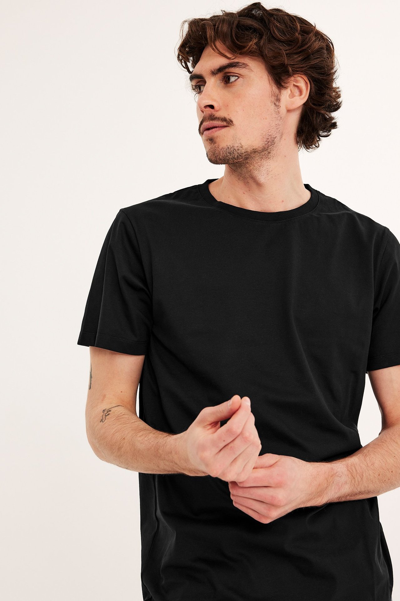 Widok z przodu i boku mężczyzny w czarnym, jednolitym, bawełnianym T-shircie z krótkim rękawem, dłonie splecione.