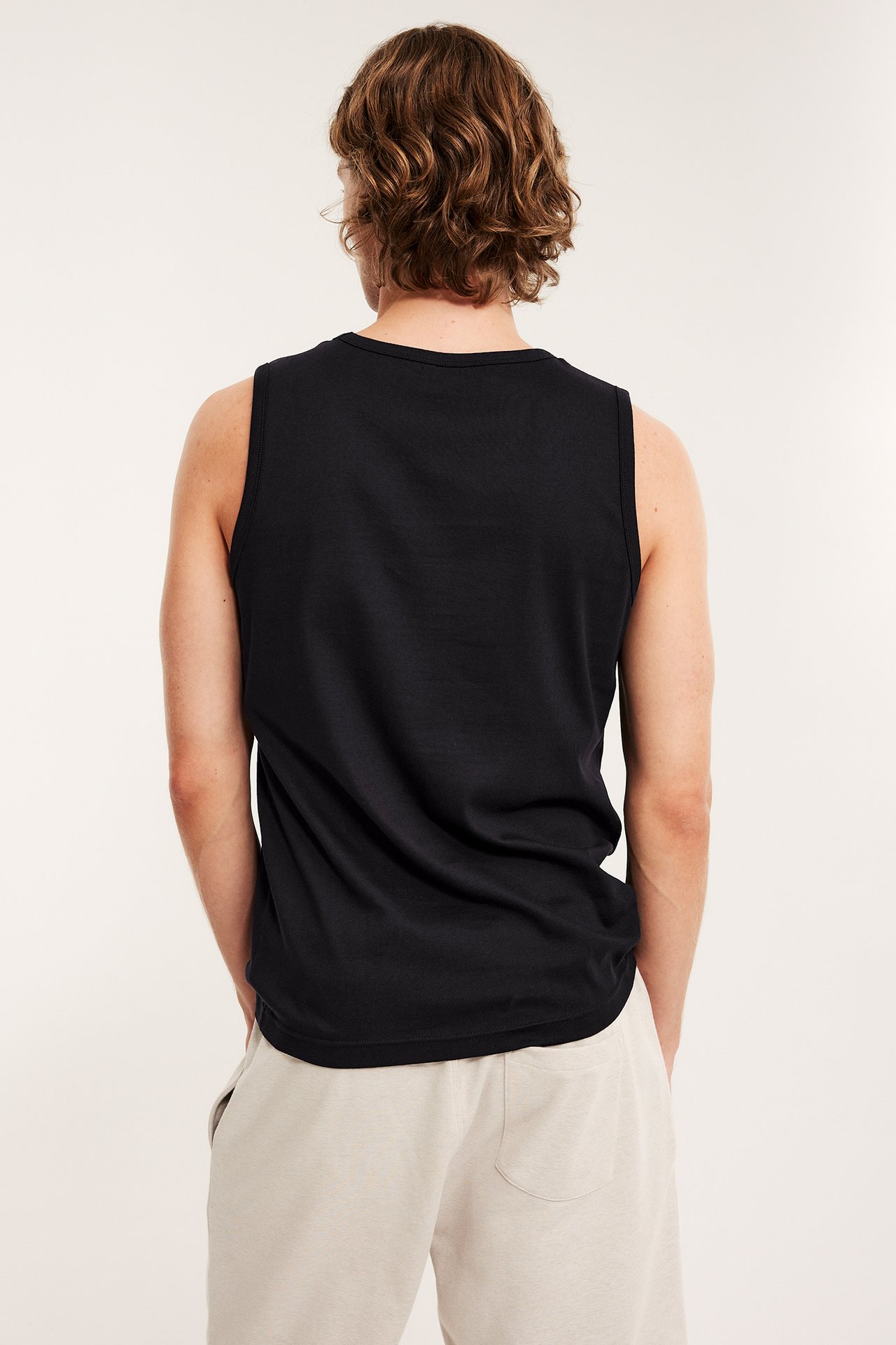 Miehen selkäkuva, yllään musta, yksivärinen puuvillainen hihaton paita ja vaaleanbeiget shortsit.