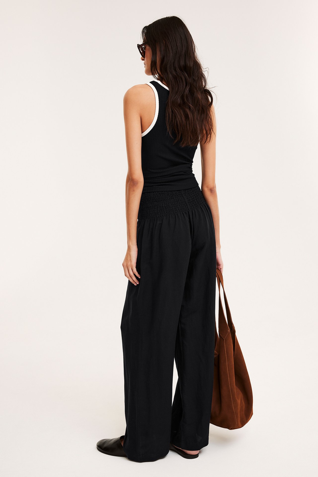 Nainen takaa: mustat leveät pellavasekoitehousut rypytetyllä vyötäröllä, musta ribbitoppi, pitää ruskeaa laukkua.