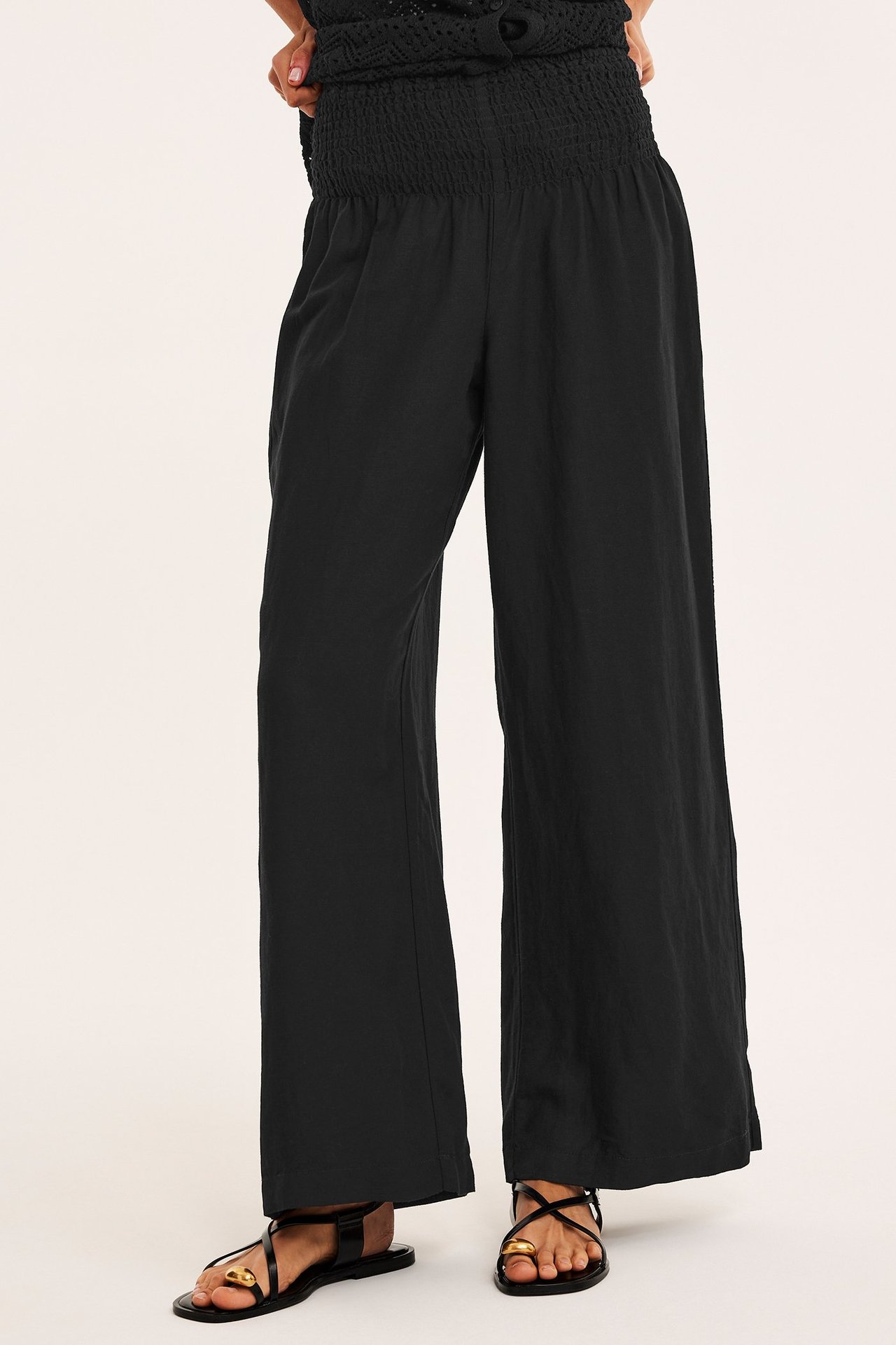 Edestä: nainen yllään mustat leveät pellavasekoitehousut rypytetyllä vyötäröllä, mustat sandaalit ja musta toppi.