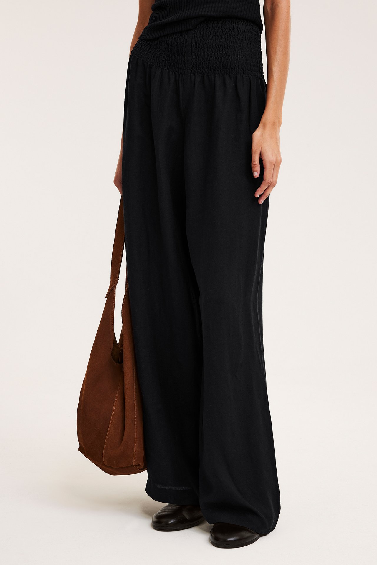 Widok z boku: Kobieta w czarnych szerokich lnianych spodniach z marszczonym pasem, czarny top, brązowa torba.