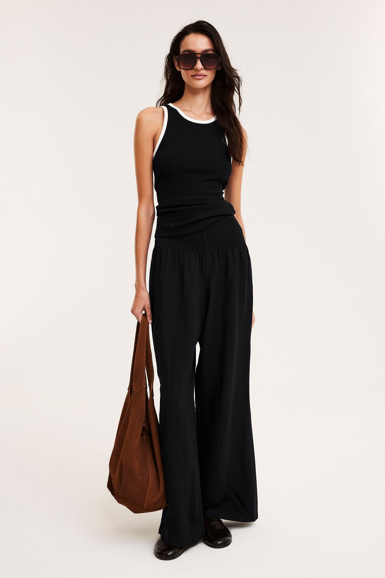 Edestäpäin: Nainen mustissa leveissä pellavahousuissa, musta ribbitoppi, aurinkolasit, pitelee ruskeaa laukkua.