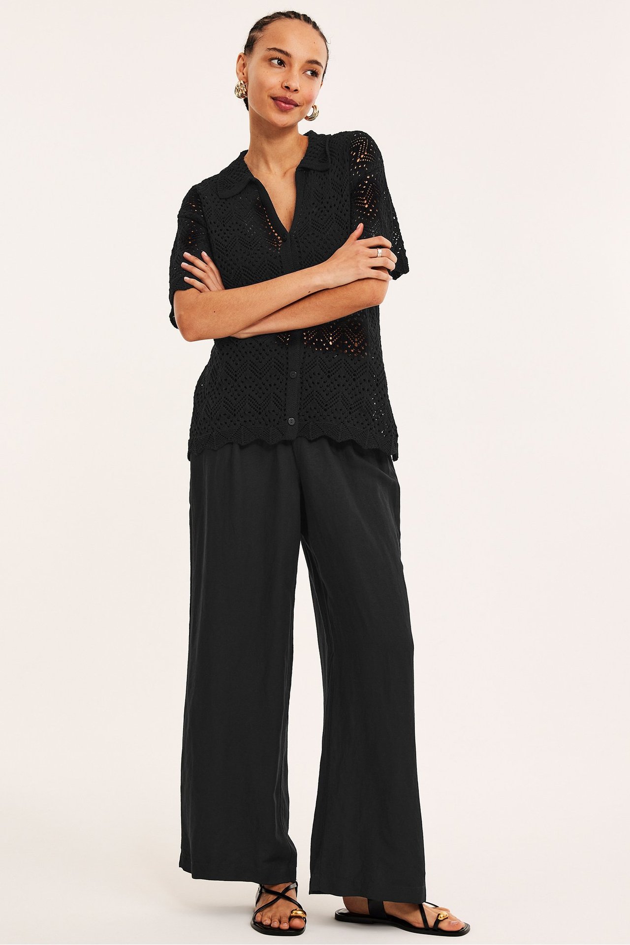 Edestä: Nainen mustissa leveissä pellavasekoitehousuissa, mustassa neulotussa topissa ja mustissa sandaaleissa.