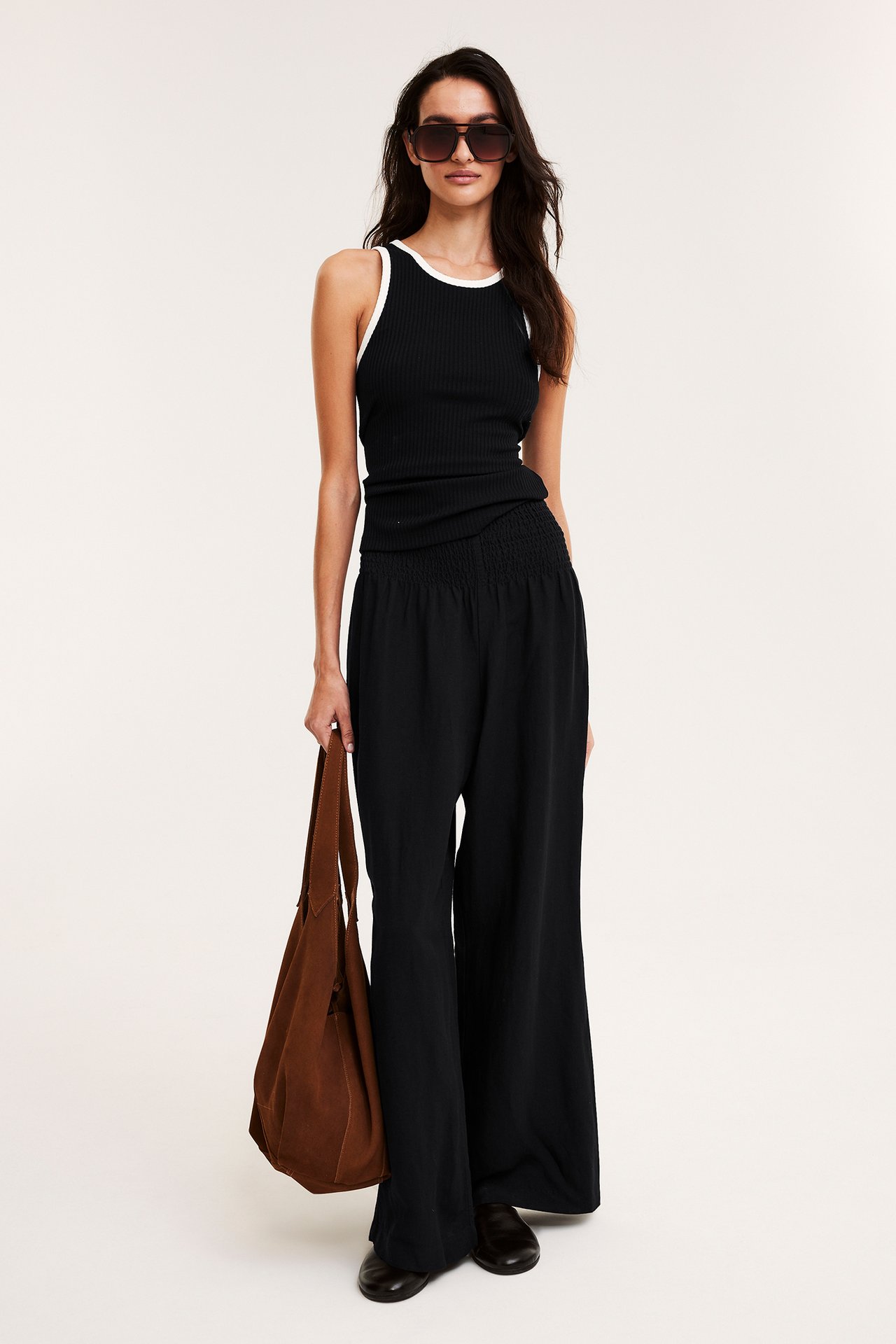 Framifrån: Kvinna i svarta vida linnebyxor, svart ribbat linne, solglasögon, håller brun väska.