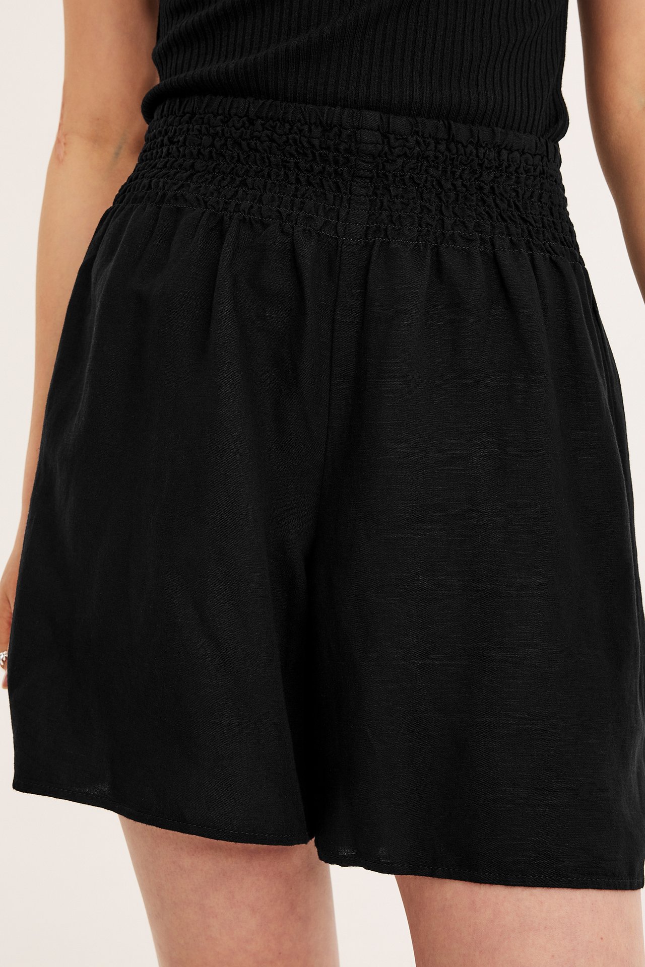 Forfra: Kvinne har på seg svarte linblandingsshorts med smockmidje og en svart ribbet topp.