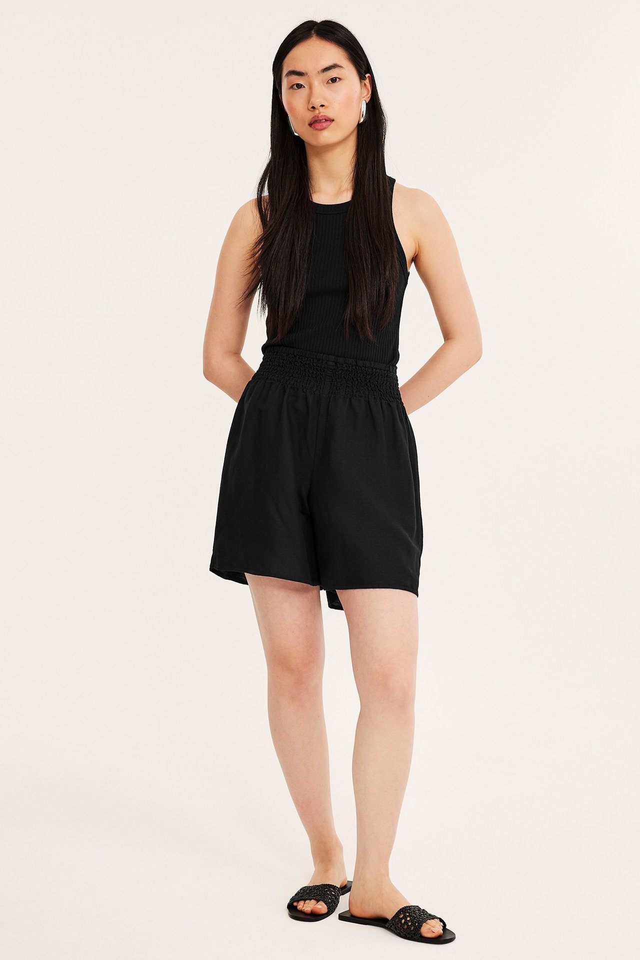 Nainen edestäpäin mustissa pellavashortseissa, joissa smokkirypytys, mustassa ribbitopissa ja mustissa sandaaleissa.