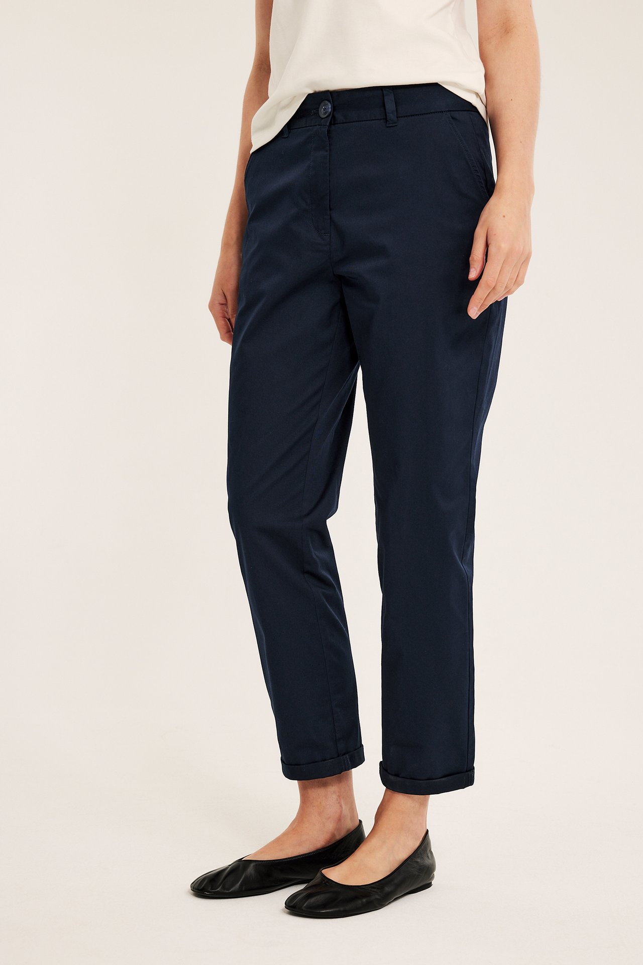 Sidovy av kvinna i mörkblå stretch-chinos med uppvikta benslut, en ljus t-shirt och svarta ballerinaskor.