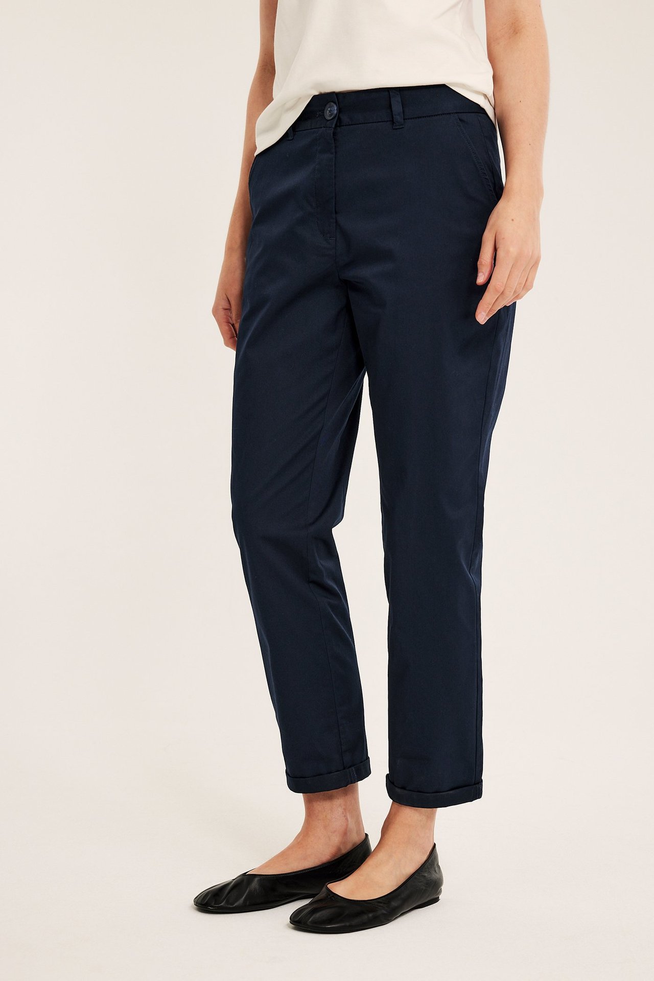 Nainen sivukuvassa tummansinisissä joustavissa puuvillachinoseissa, käärityt lahkeet, vaalea t-paita ja mustat ballerinat.