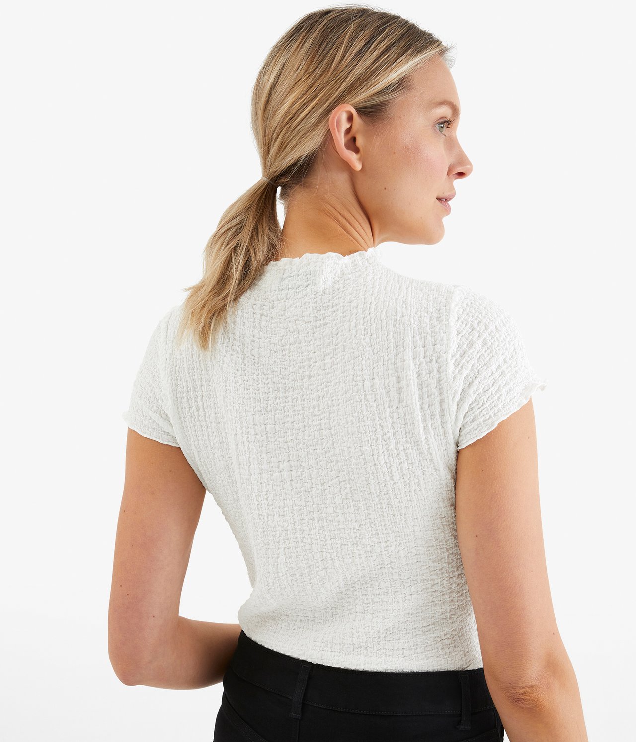 Sned bakifrån-vy av en kvinna i en offwhite texturerad, figurnära kortärmad T-shirt och svarta byxor.
