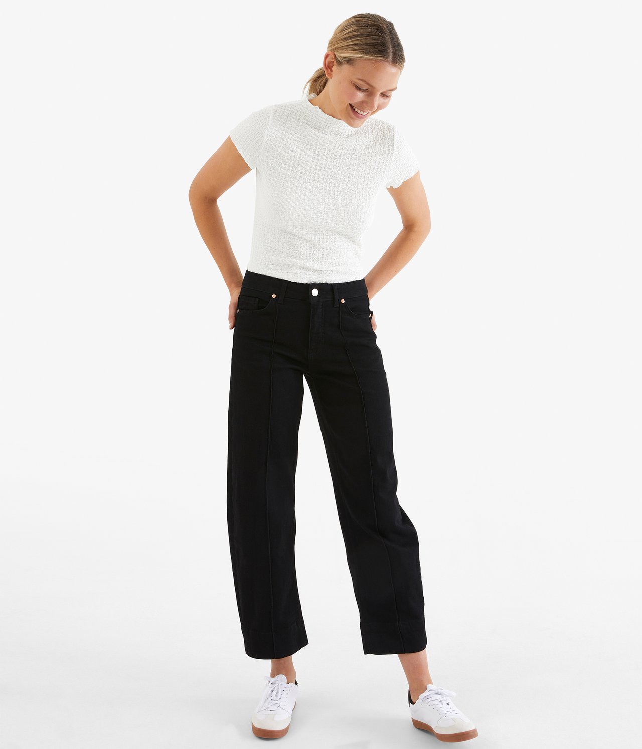 Framifrån: kvinna i offwhite texturerad åtsittande t-shirt, svarta vida croppade jeans och vita sneakers.