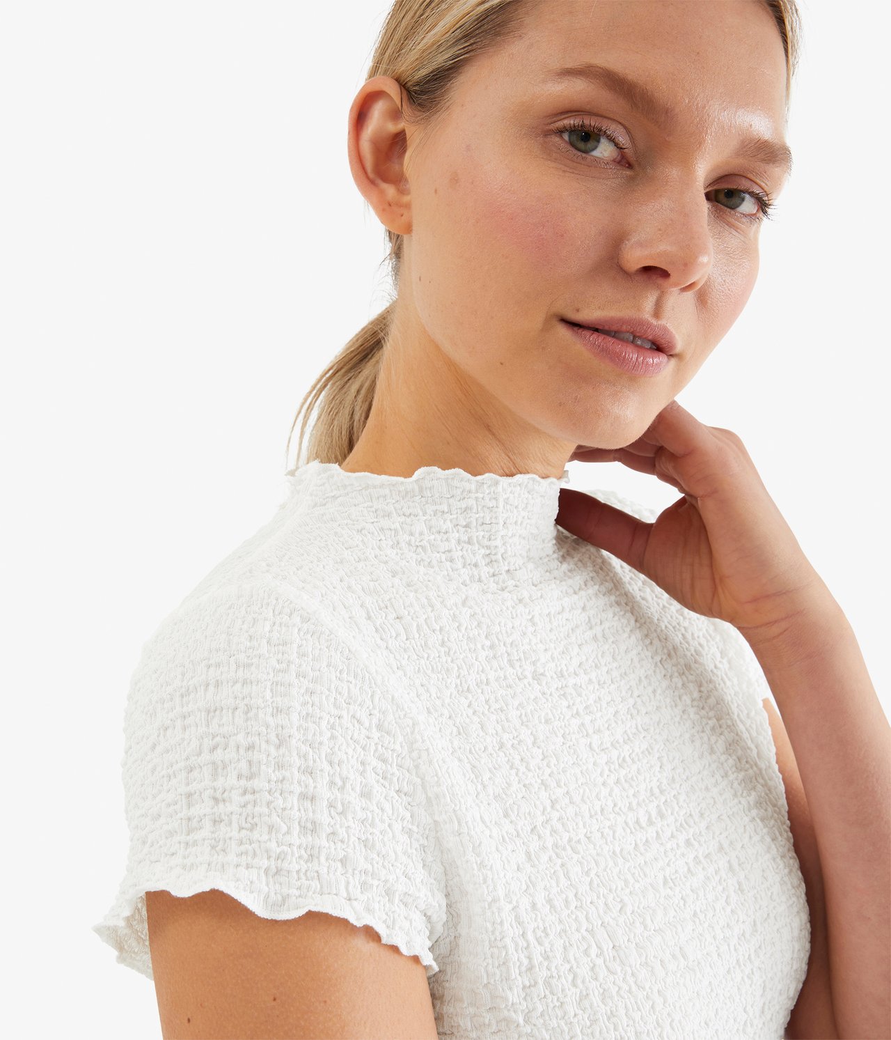 Närbild på en kvinna i en offwhite texturerad figurnära T-shirt med volanghals och korta ärmar.