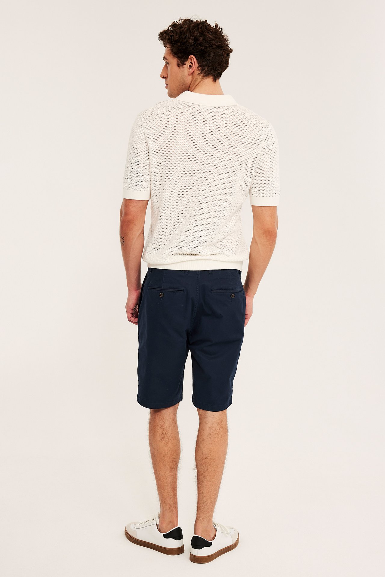 Bakifrån på man i mörkblå bomullstwill chinosshorts, vit stickad pikétröja och vita sneakers.