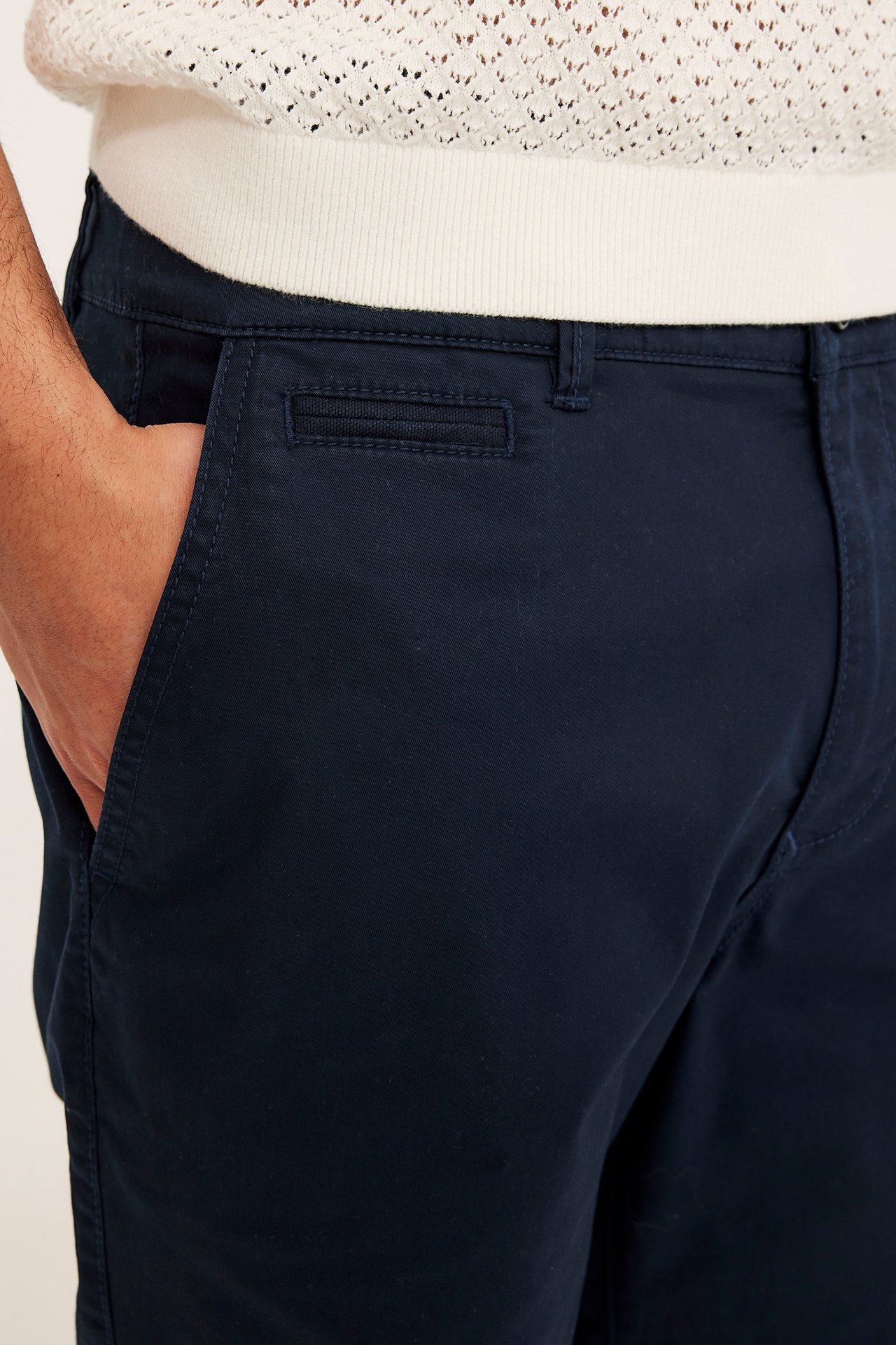Zbliżenie mężczyzny w granatowych bawełnianych szortach chino, ręka w kieszeni, na białym swetrze.