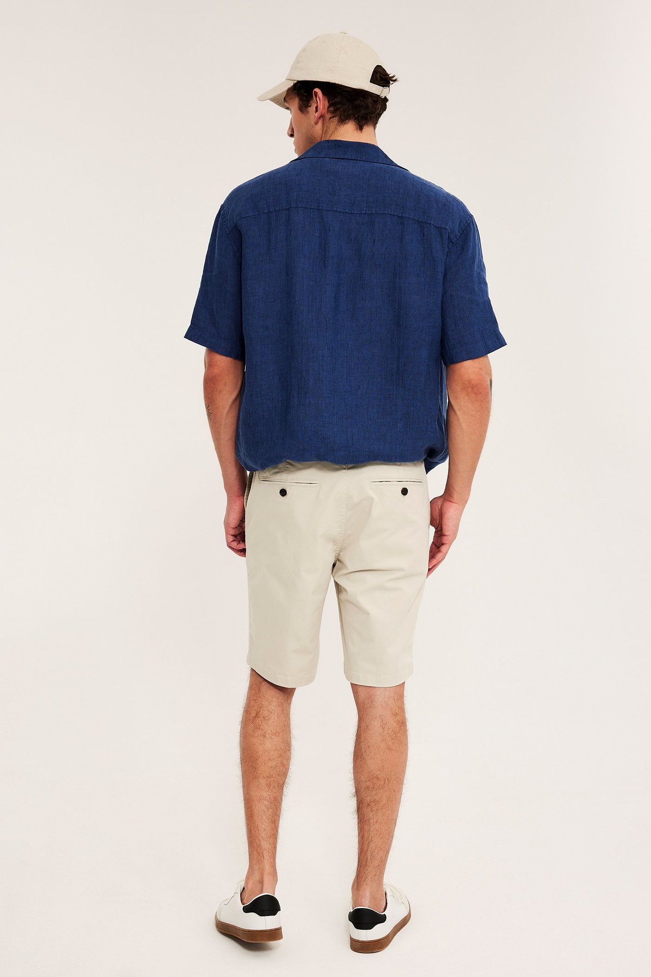 Widok z tyłu: mężczyzna w jasnobeżowych szortach chinos, niebieskiej koszuli, beżowej czapce i białych sneakersach.