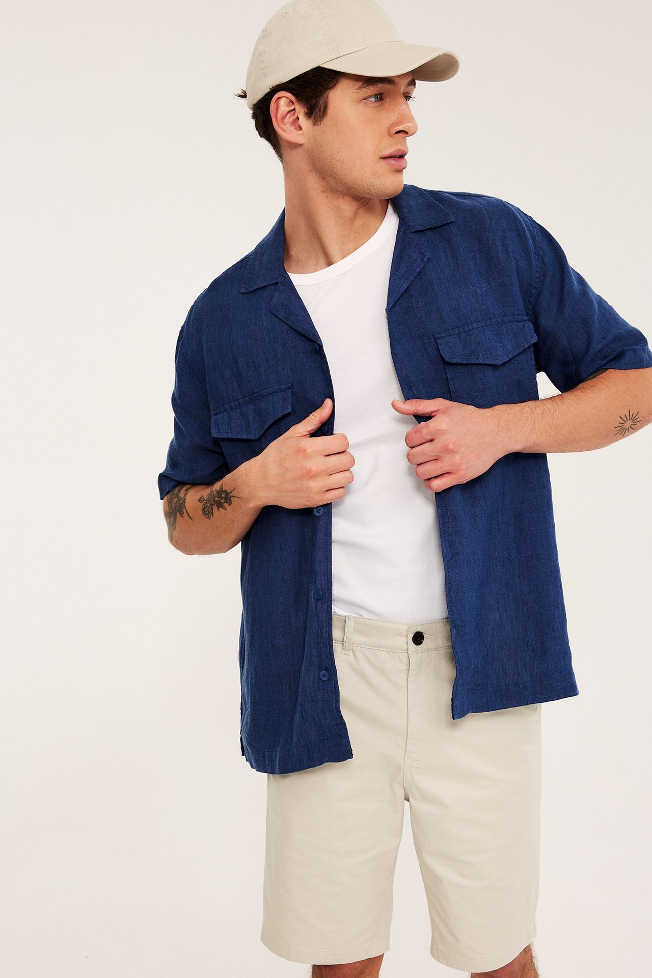 Mies edestäpäin vaaleanbeigissä chino-shortseissa, sinisessä avoimessa paidassa, valkoisessa t-paidassa ja beigessä lippalakissa.