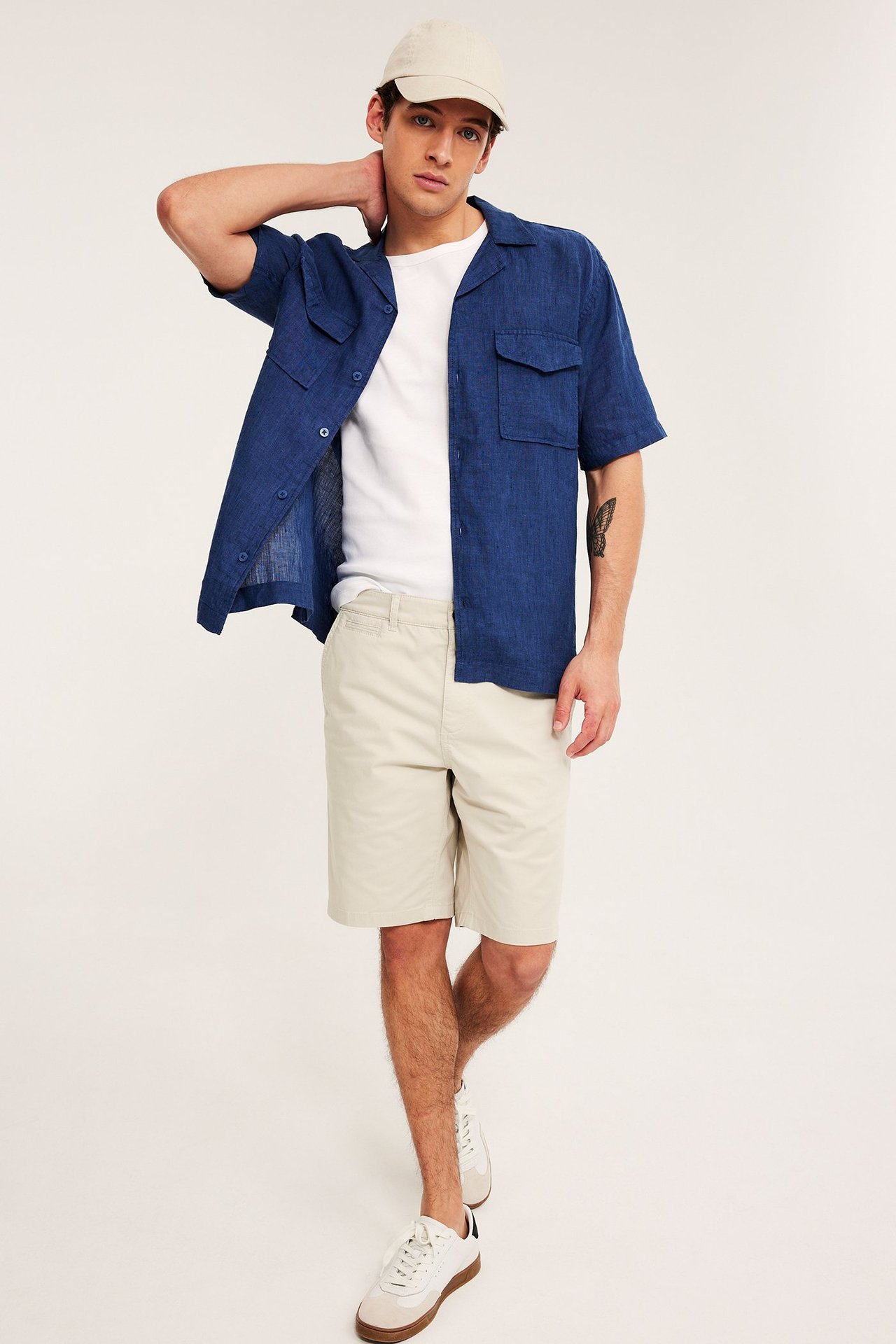Front view: Man i ljusbeige chinoshorts, vit t-shirt, öppen blå skjorta, beige keps, vita sneakers.