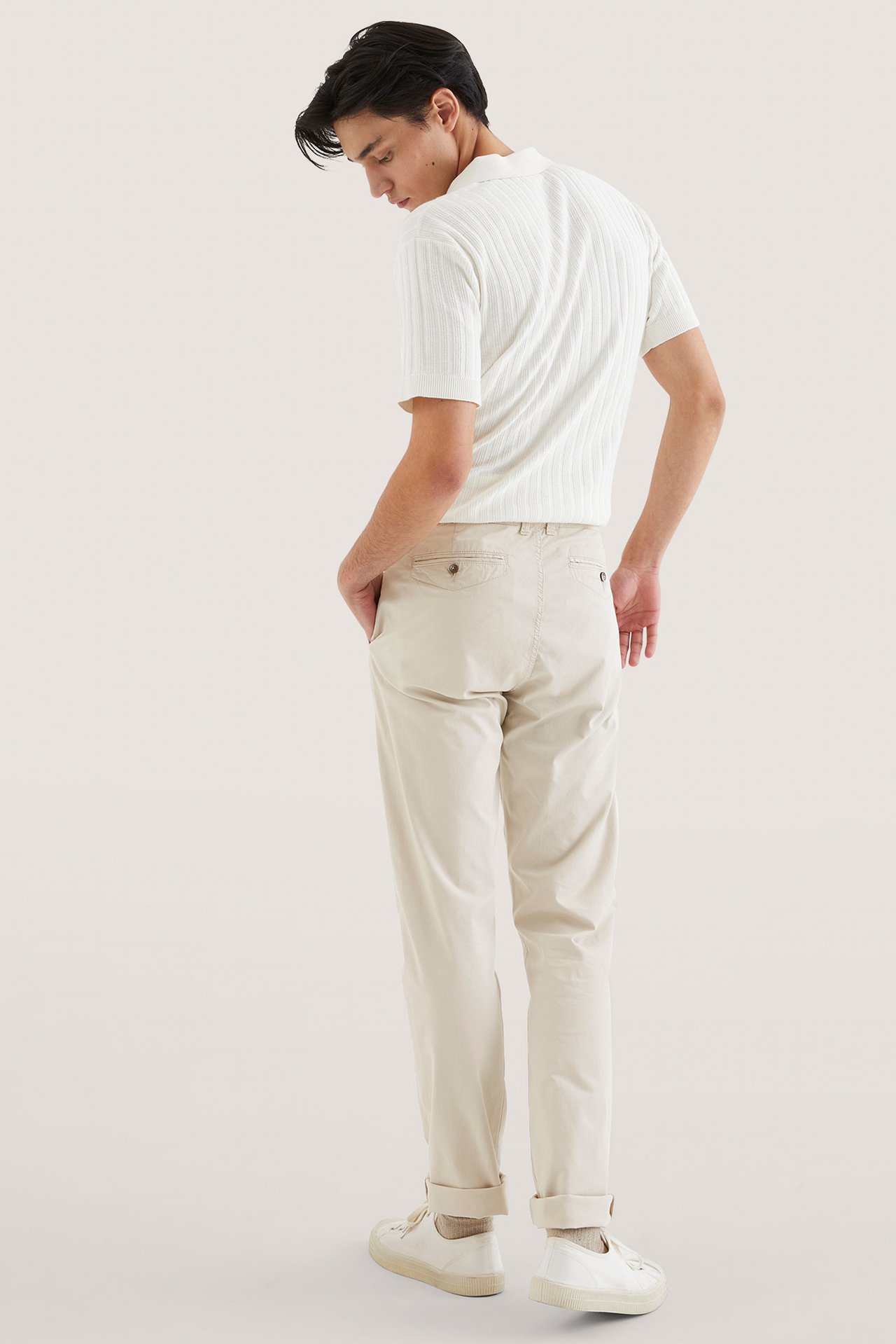 Bakfra, mann i lysebeige regular fit chinos, hvit ribbet pikétrøye og hvite joggesko.