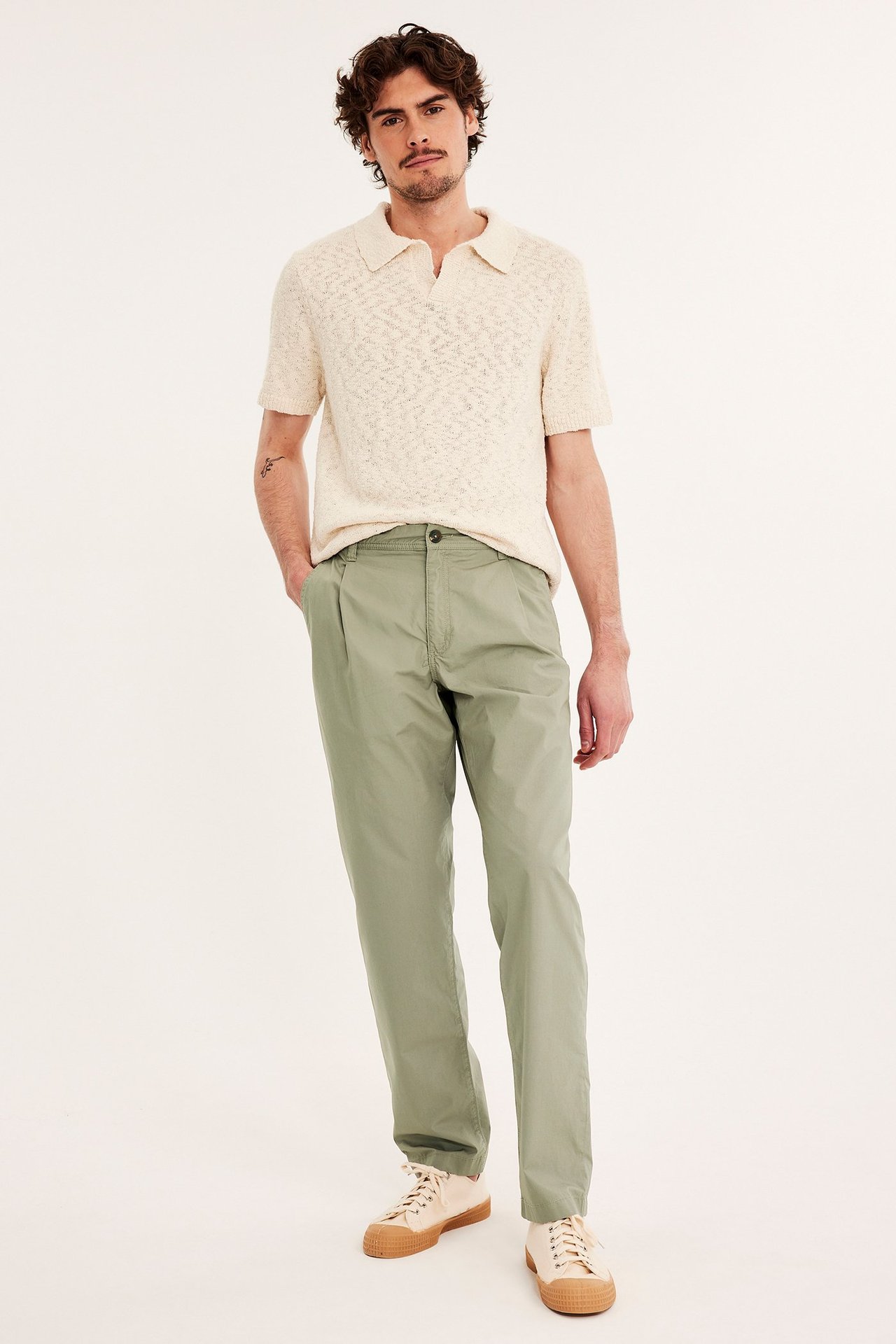 Edestä kuvattu mies vihreissä laskostetuissa chinoissa, beigessä neulotussa pikeepaidassa ja beigissä tennareissa.