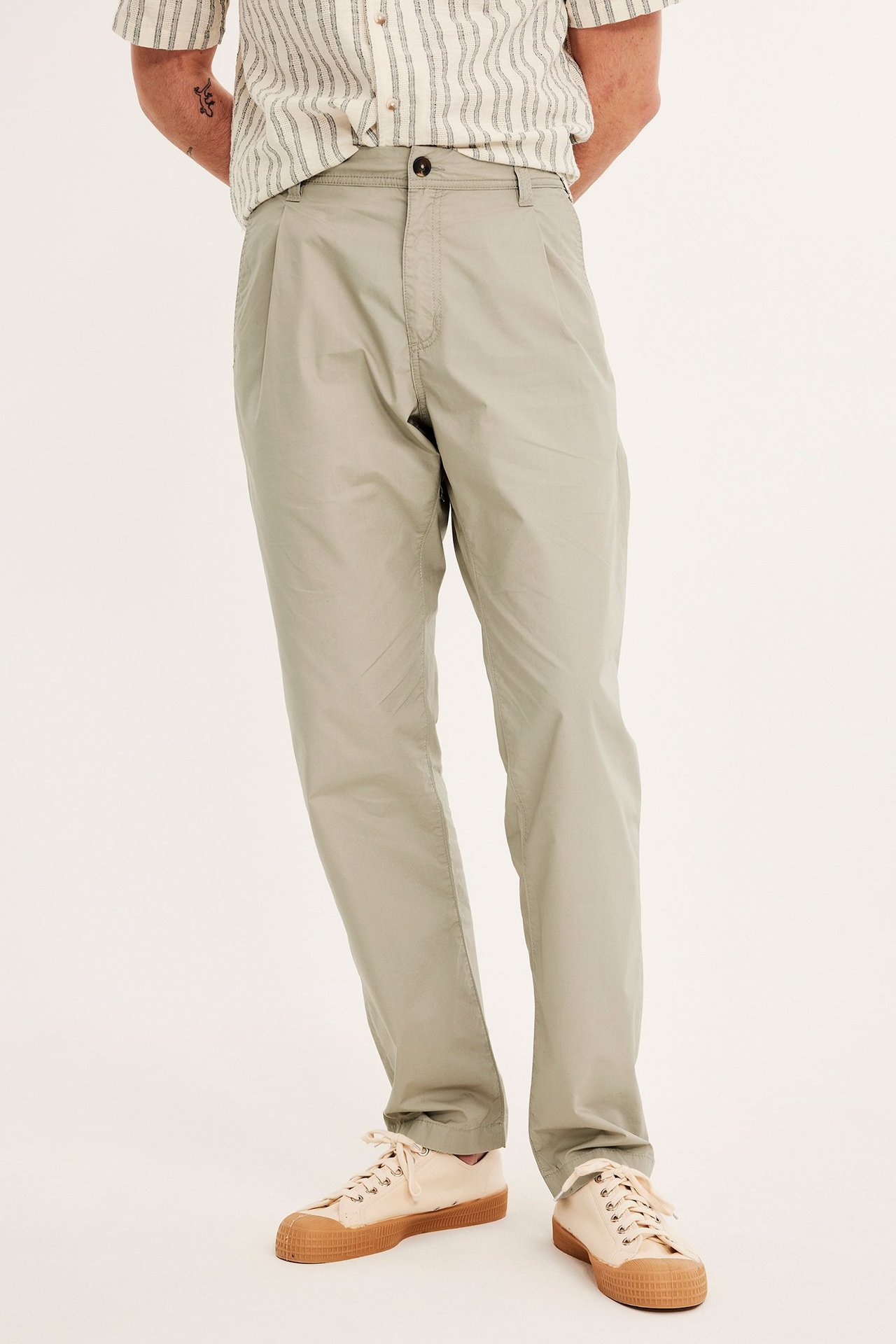 Edestä kuvattu mies beigessä laskostetussa chinohousuissa, raidallisessa lyhythihaisessa paidassa ja beigissä kangaskengissä.