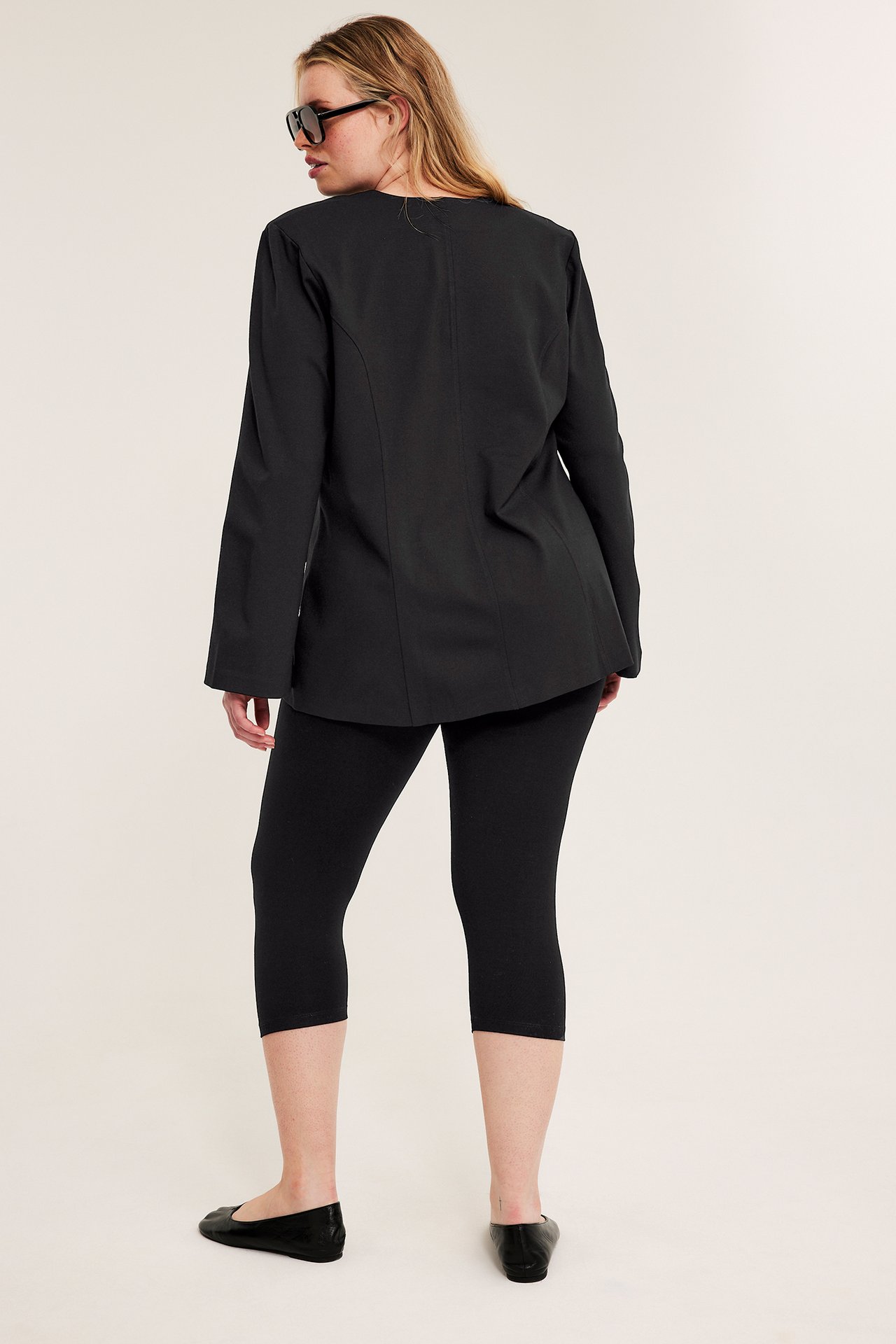 Bakifrån: Kvinna i svarta caprileggings, lång svart tunika, svarta ballerinaskor och solglasögon.
