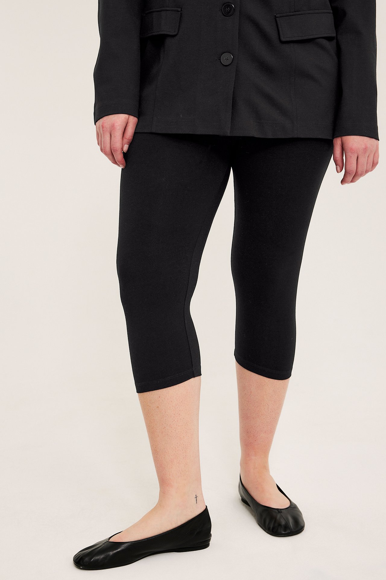 Framifrån: kvinna i åtsittande svarta caprileggings, en svart kavaj och svarta ballerinaskor.