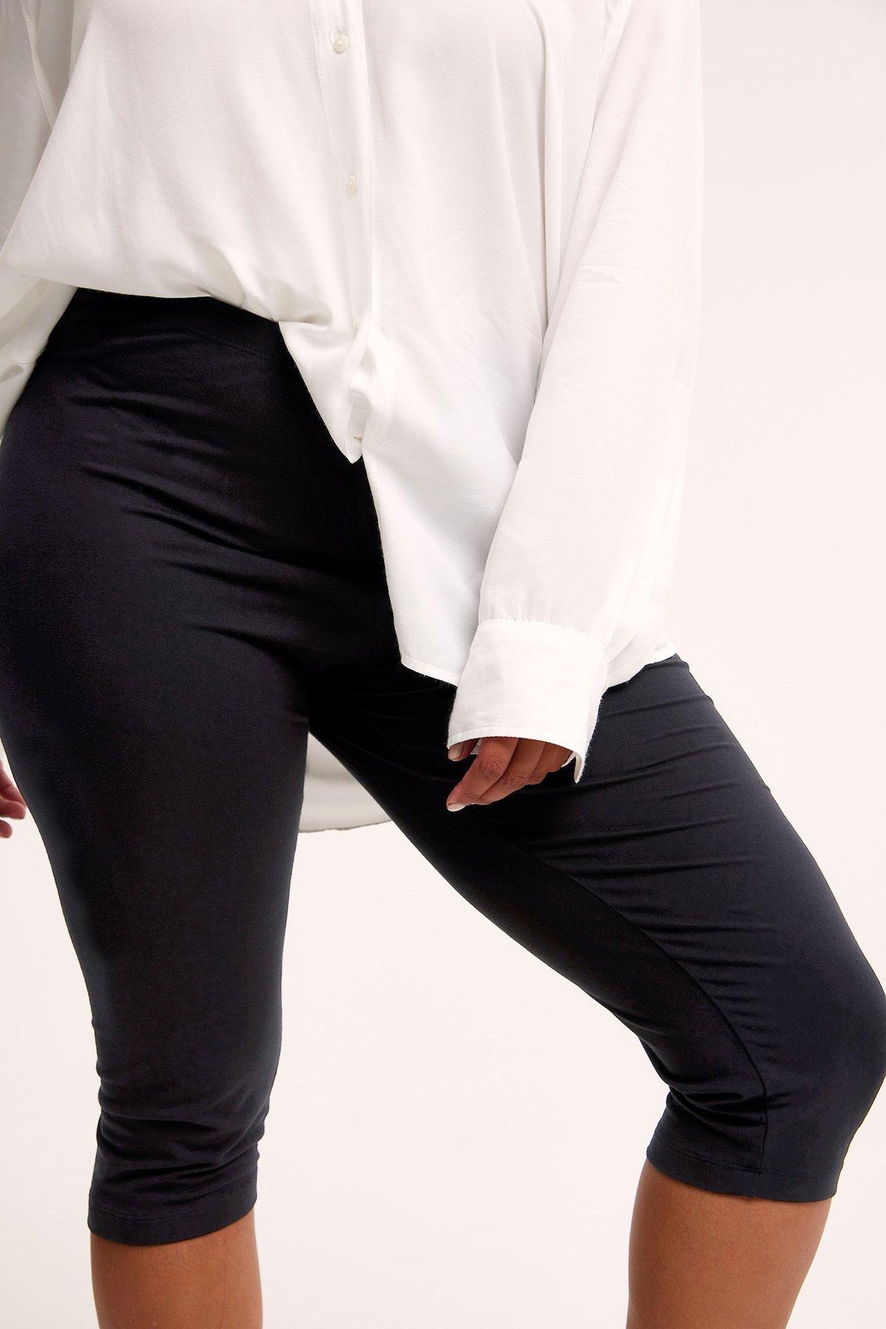 Edestäpäin, nainen yllään mustat vartalonmyötäiset capri-leggingsit ja väljä valkoinen pitkähihainen paita.