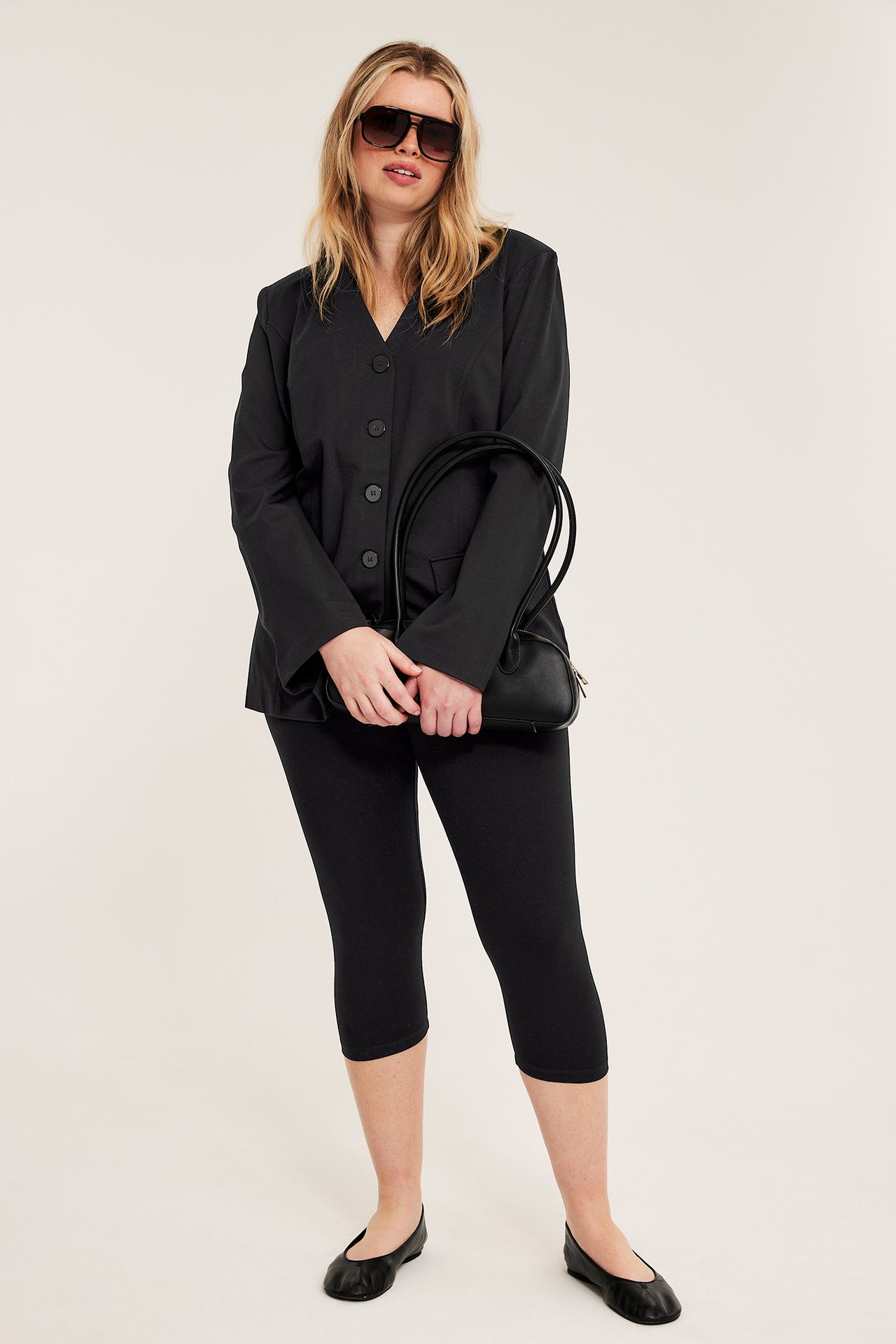 Frontutsikt: Kvinne i svarte caprileggings, svart blazer, flate sko, solbriller, holder en svart håndveske.