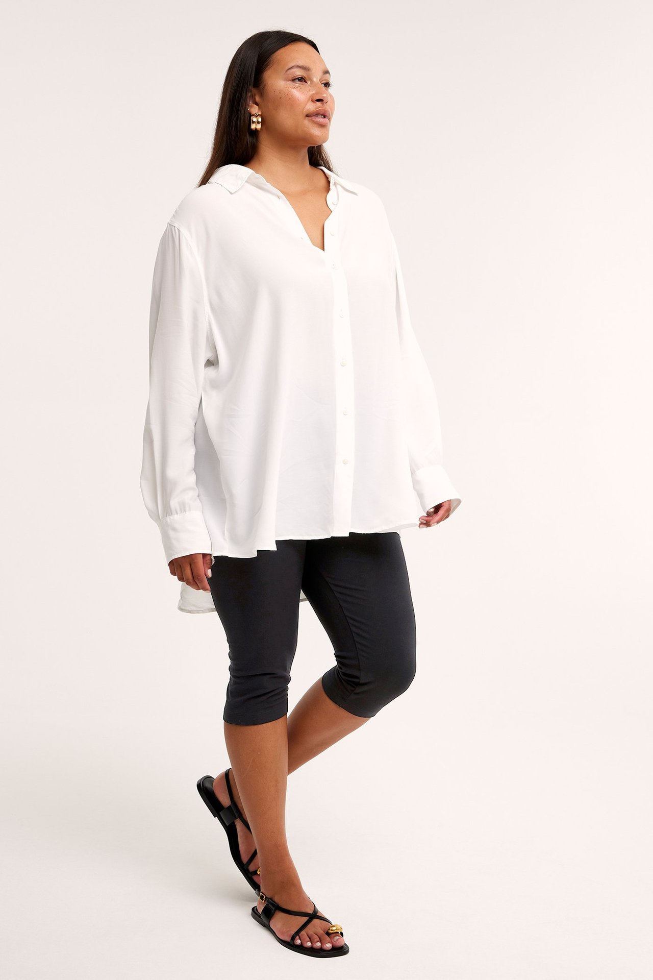 Widok z boku kobiety w czarnych legginsach capri, białej koszuli, czarnych sandałach i złotych kolczykach.
