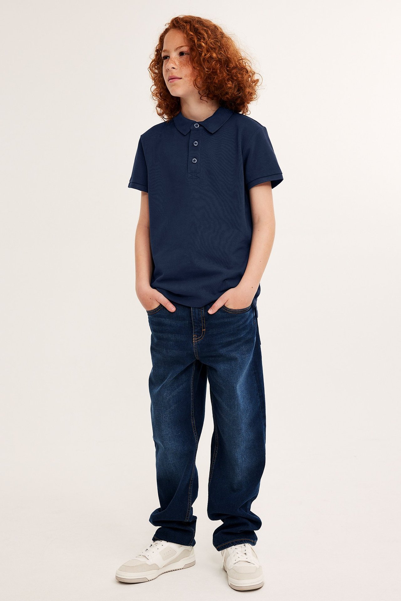 Frontvy av ett barn i mörkblå bomullspikétröja med knappar, mörkblå jeans och vita sneakers.