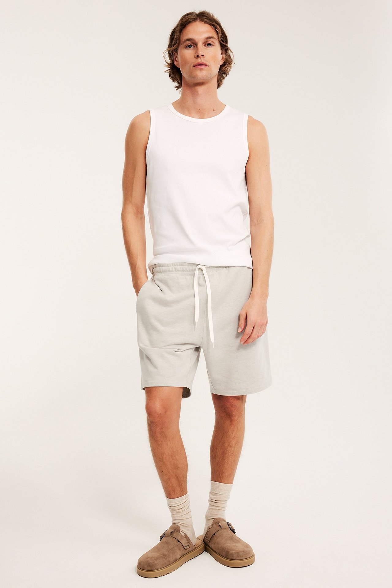 Framifrån, en man i melerade beiga mjukisshorts med dragsko, vitt linne och bruna tofflor.