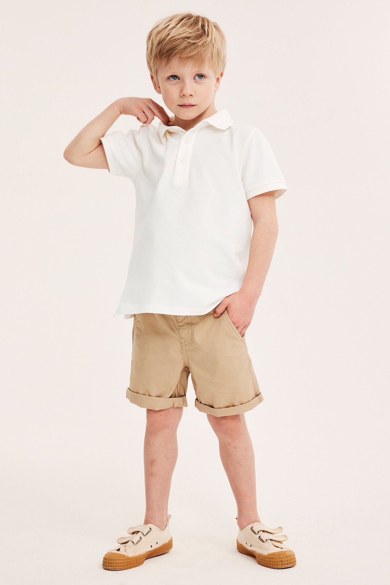 Frontvy av ett barn i en offwhite pikétröja i bomull, beiga shorts och ljusbeiga sneakers.