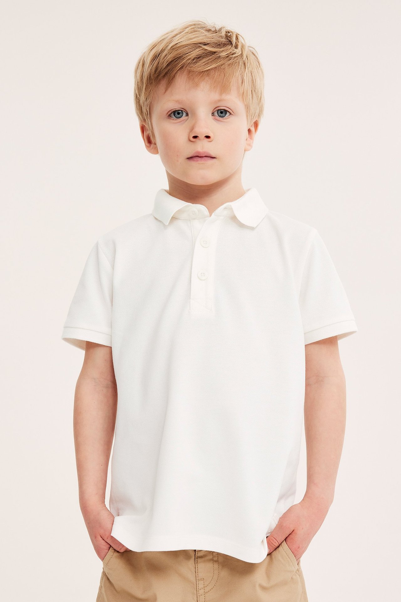 Frontvy av ett barn i en offwhite pikétröja i bomull med knappar, matchad med beiga shorts.