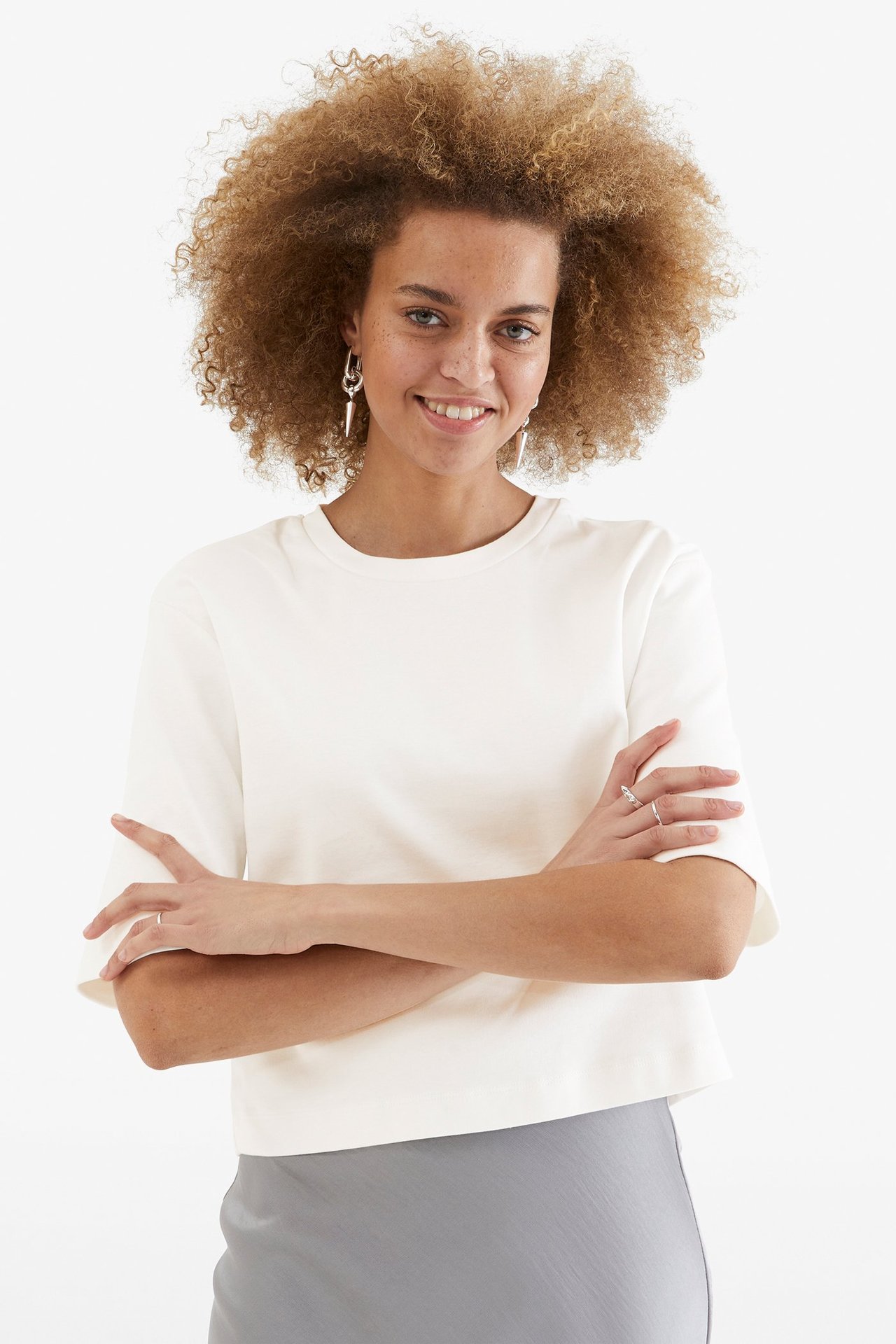 Kobieta w kremowym, krótkim bawełnianym t-shircie, szarej spódnicy, srebrnych kolczykach i pierścionkach.