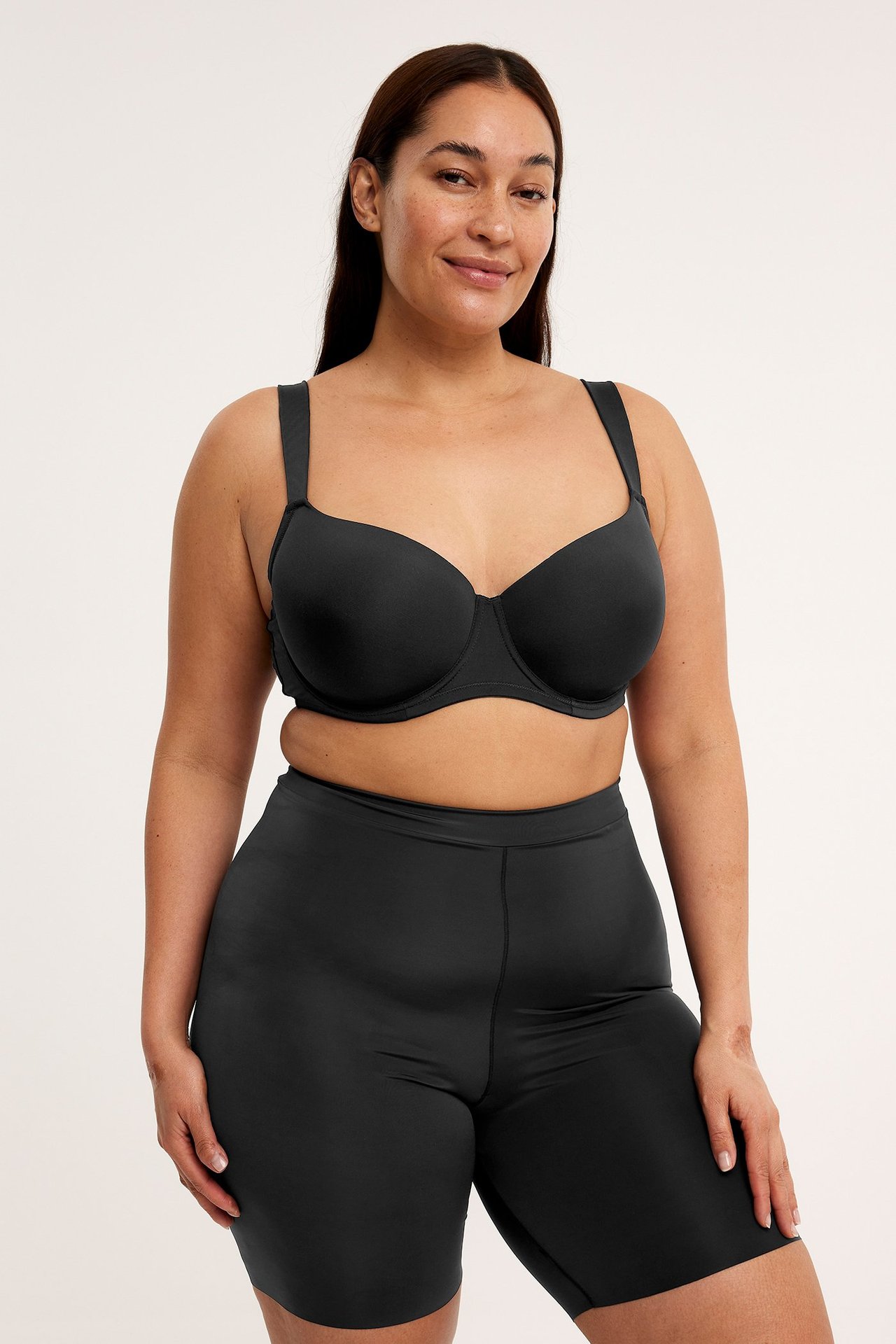 Forfra, kvinne i svarte, ensfargede shaping-shorts av elastisk stoff, sammen med en svart BH.