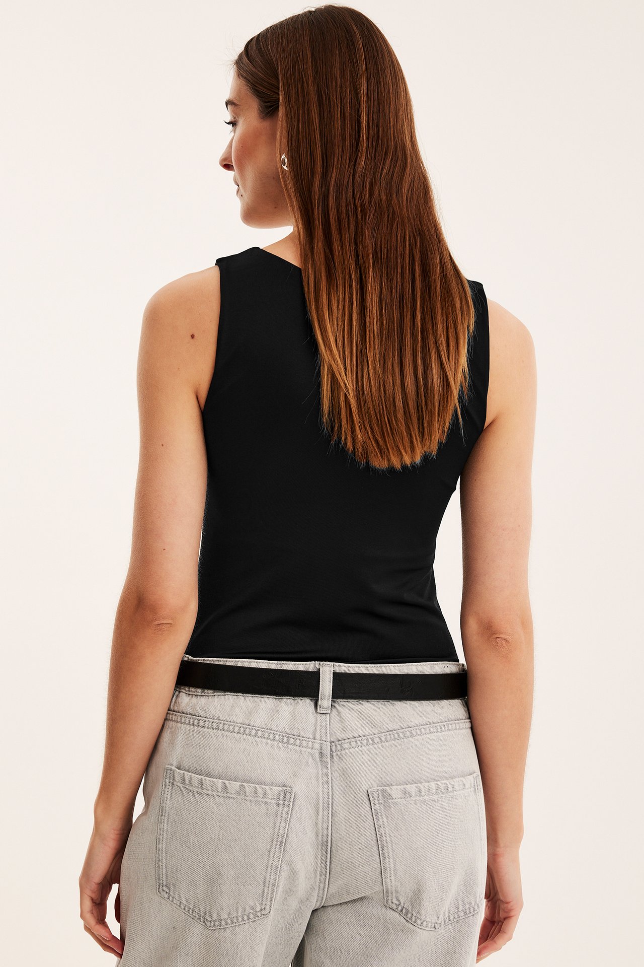 Bakifrån: en kvinna i ett svart tight ärmlöst linne, ljusgrå jeans och ett svart bälte.