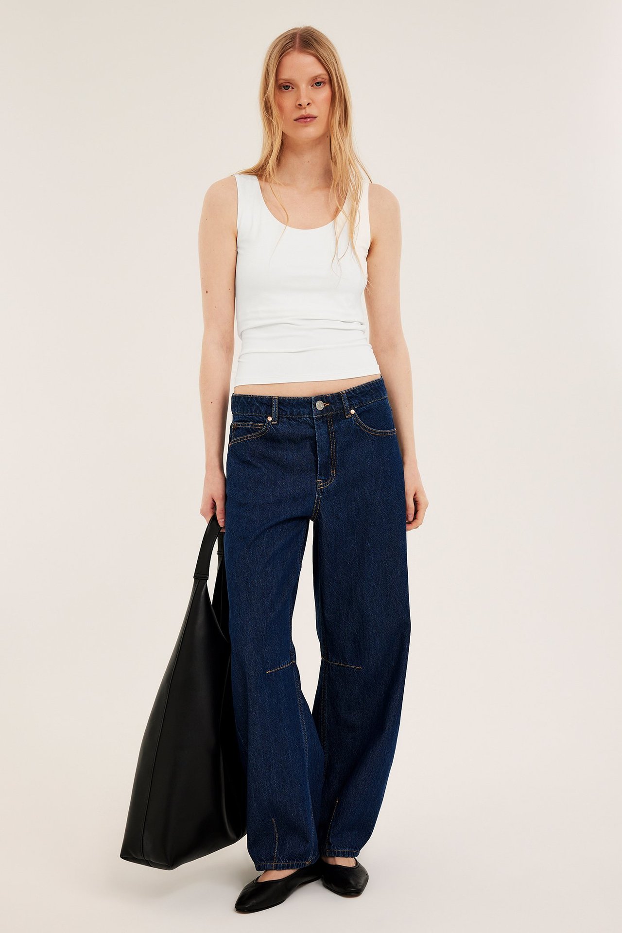 Frontvy av kvinna i offwhite tight linne, mörkblå vida jeans, svarta platta skor, håller svart tygväska.