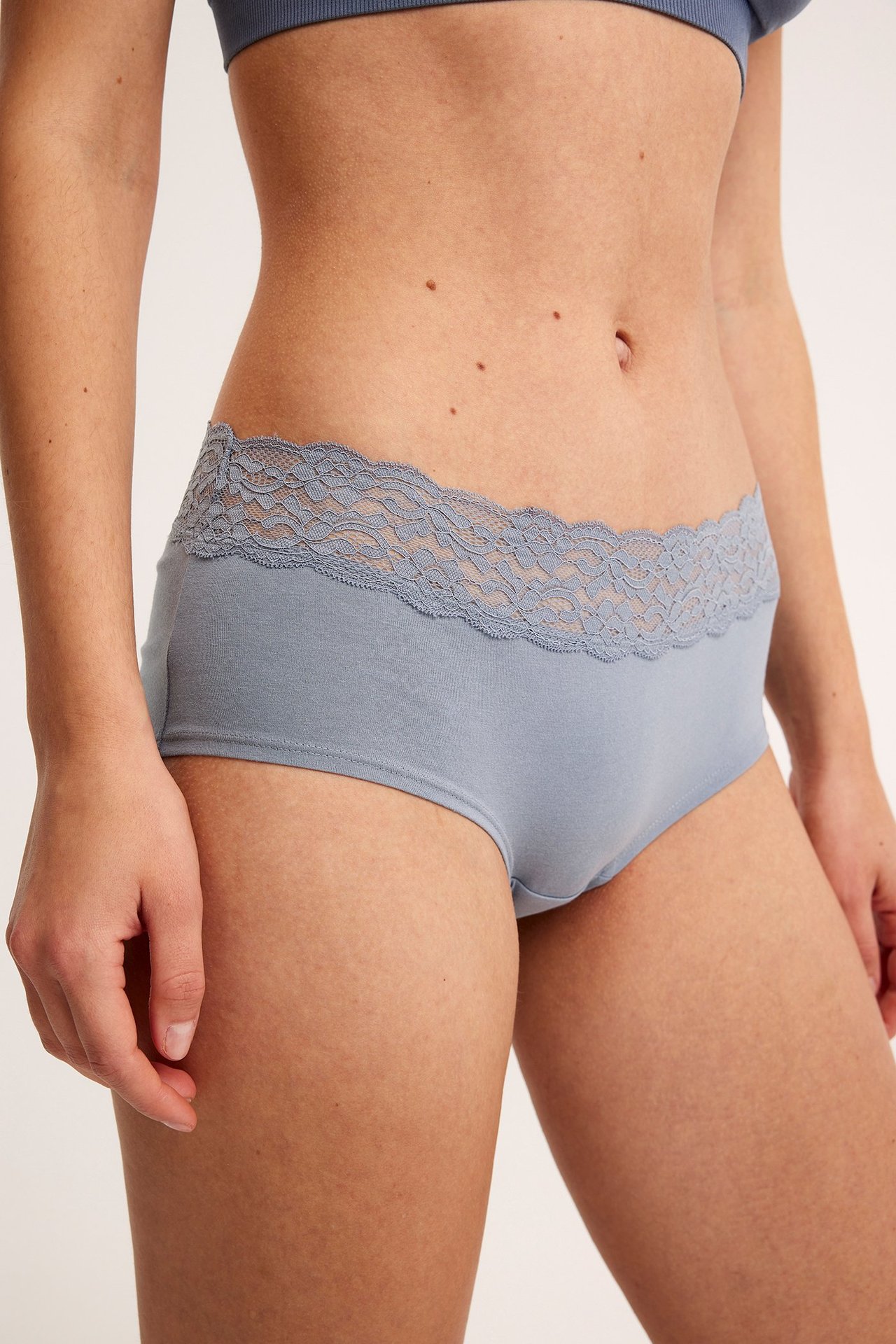 Frontvy av kvinna i ljusblå bomullshipstertrosa med spets i midjan och matchande BH.