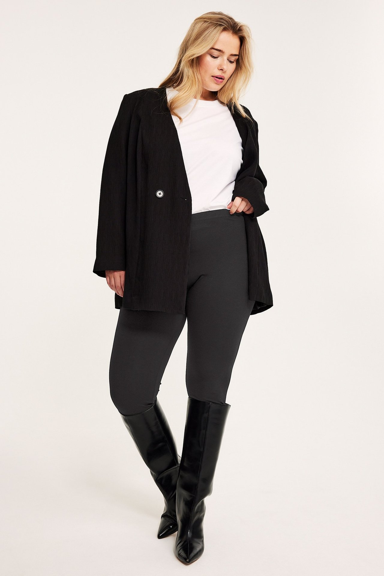 Framifrån, kvinna i svarta jersey-leggings, vit t-shirt, öppen svart kavaj och svarta knähöga stövlar.