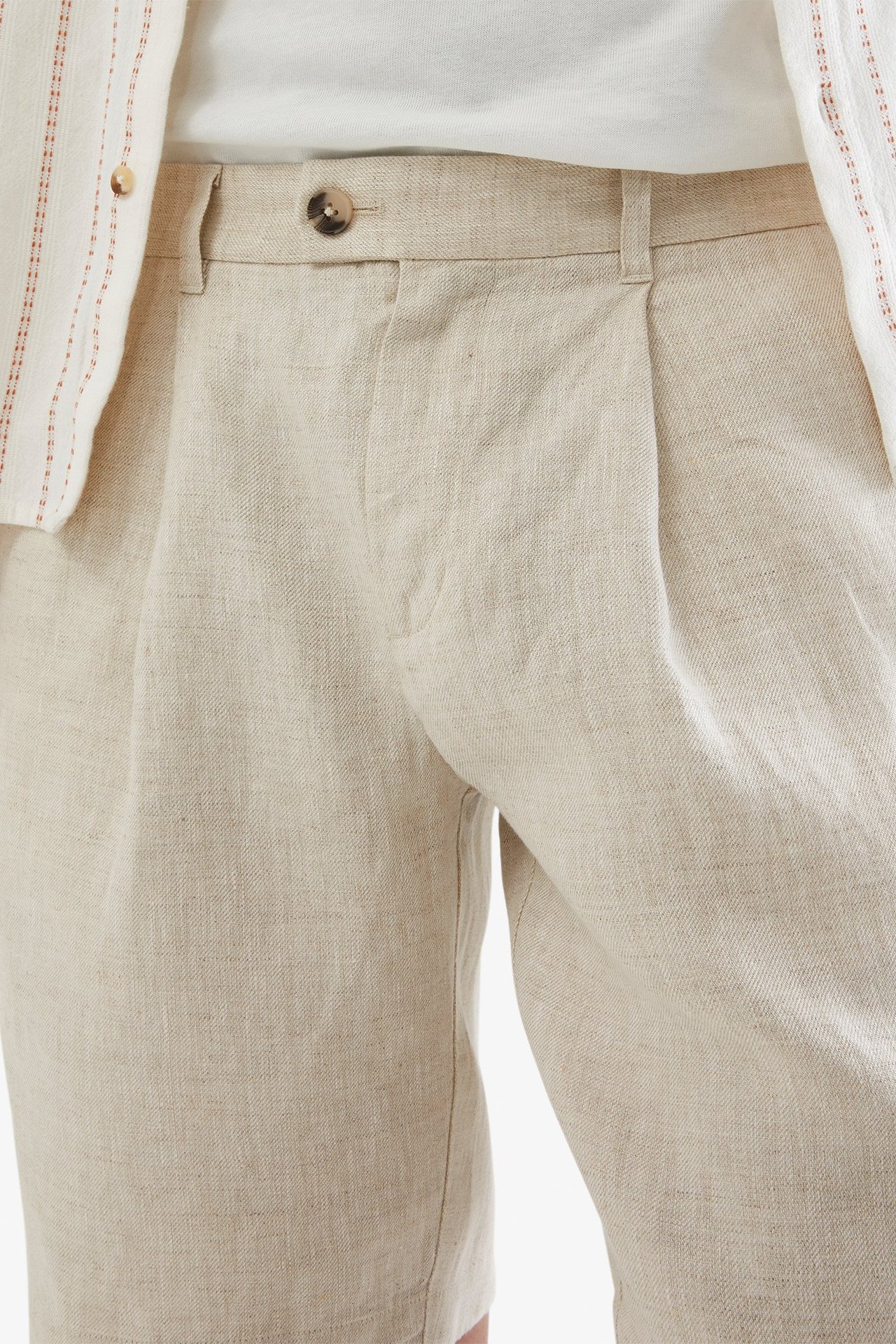 Lähikuva edestä miehestä beigessä pellavashortseissa, joissa laskos, valkoinen t-paita ja raidallinen paita.