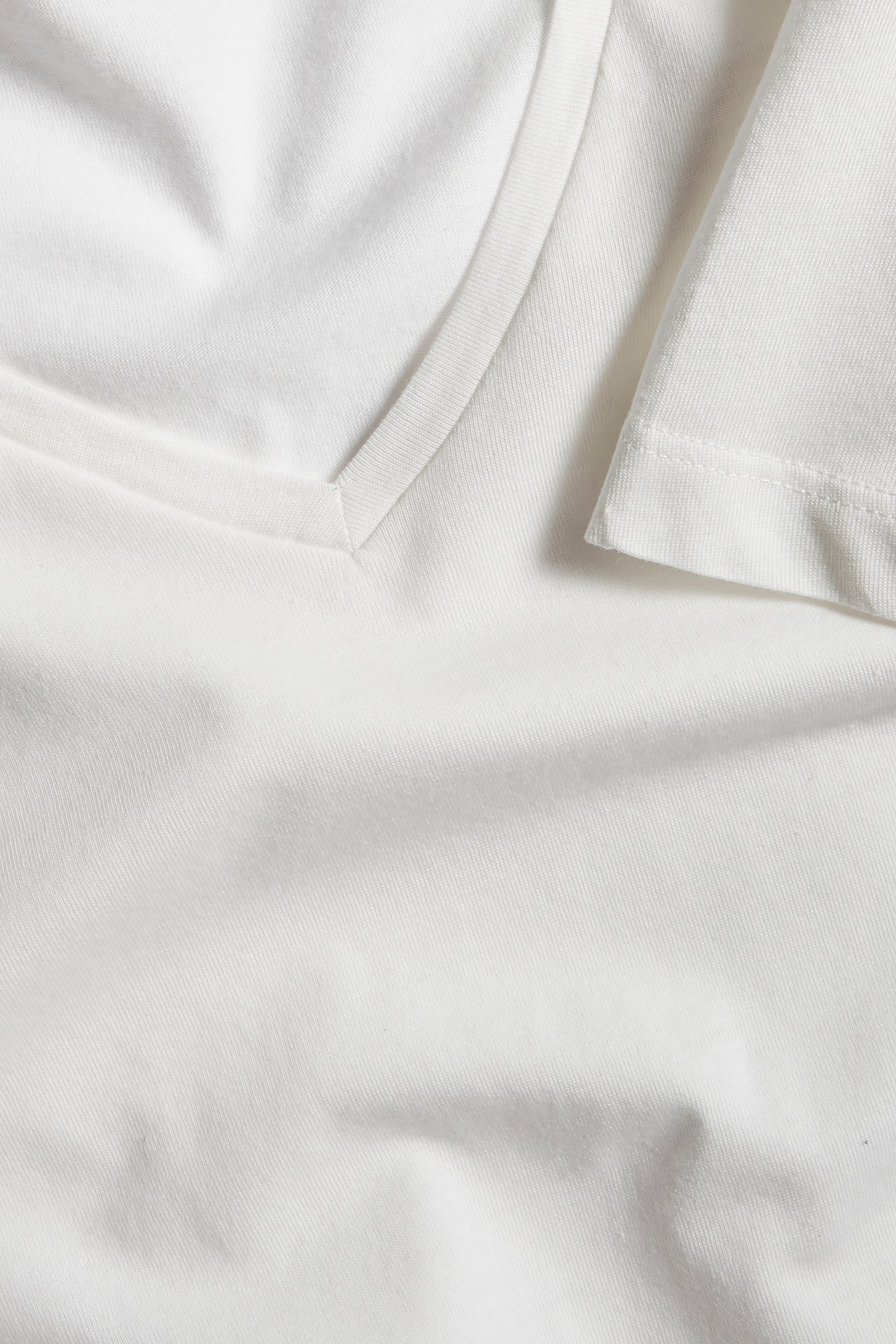 Zbliżenie na kremowy T-shirt z dekoltem w serek, wykonany z cienkiej bawełny, widoczny dekolt i brzeg rękawa.
