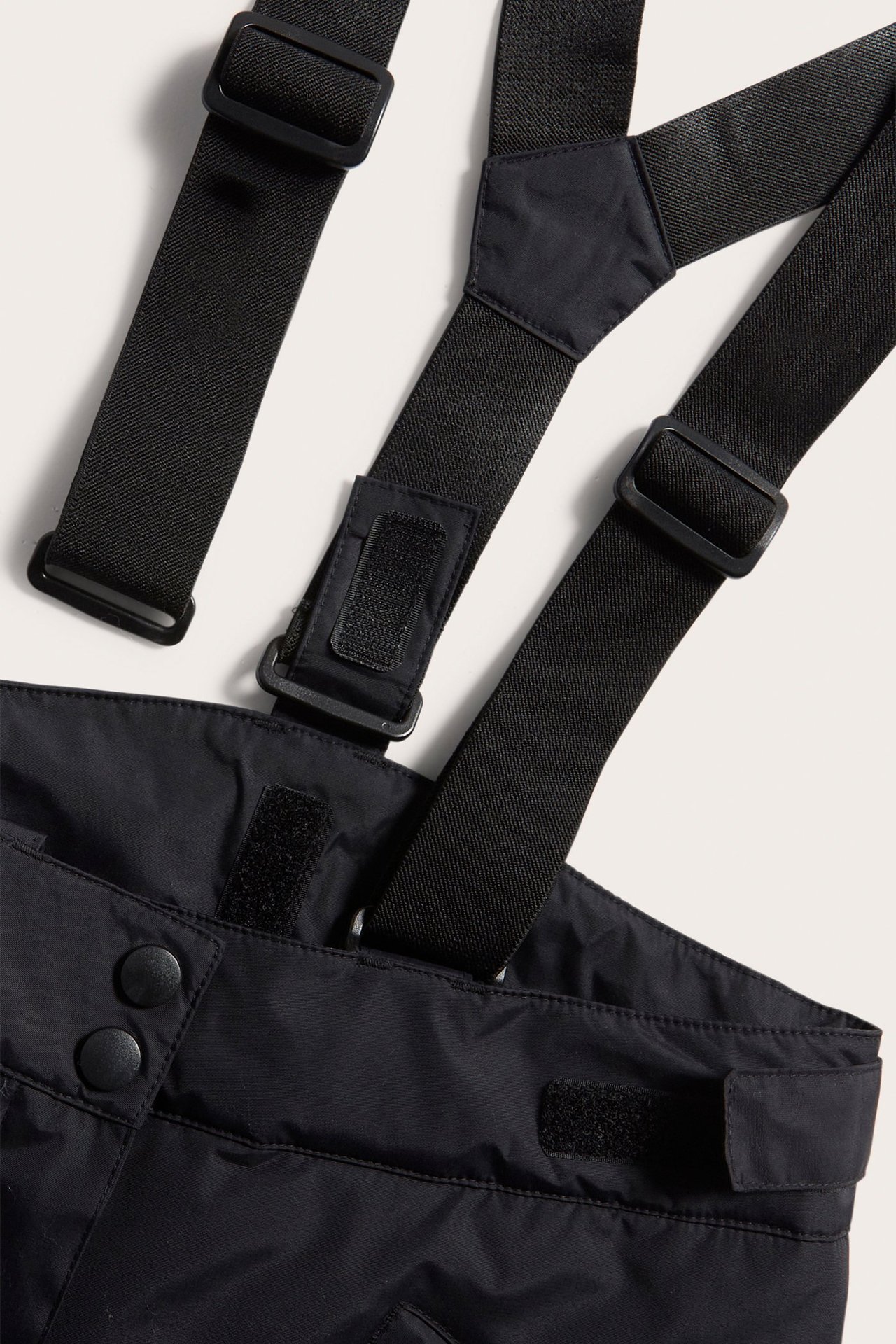 Zbliżenie czarnych spodni narciarskich z regulowanymi szelkami, klamrami, rzepami i guzikami.