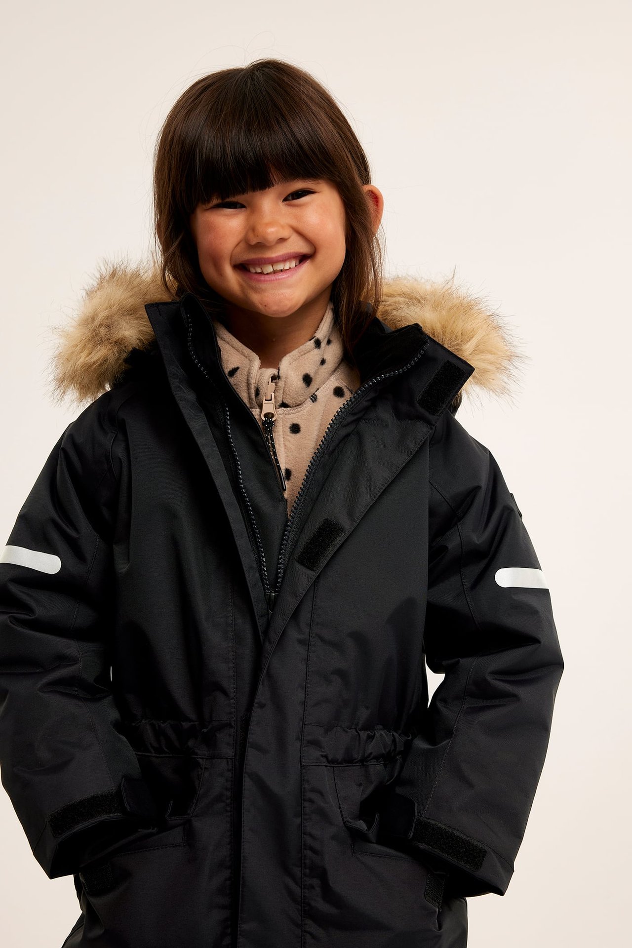 Barn i svart Kaxs Proxtec vinteroverall med fuskpäls på huvan, över en beige prickig fleece.