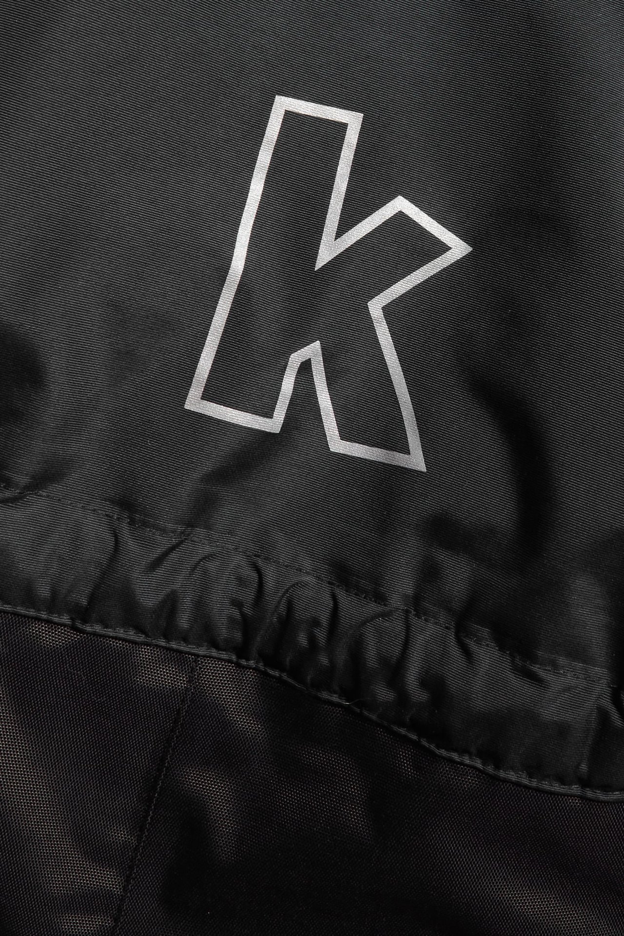 Zbliżenie czarnego kombinezonu zimowego Kaxs Proxtec z widocznym białym logo „k”.