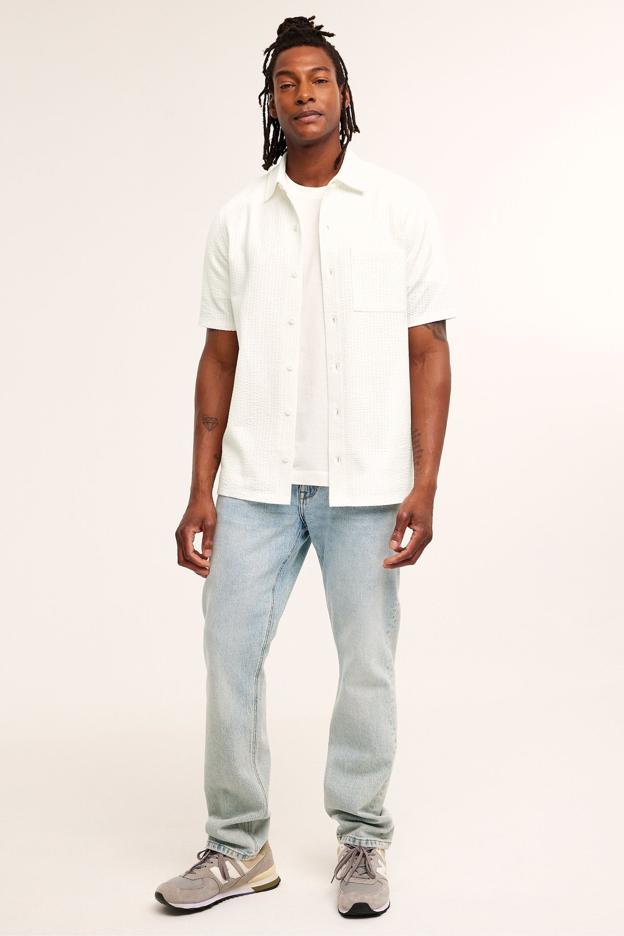 Edestä: mies luonnonvalkoisessa raidallisessa lyhythihaisessa trikoopaidassa, valkoisessa t-paidassa, vaaleansinisissä farkuissa, harmaissa lenkkareissa.