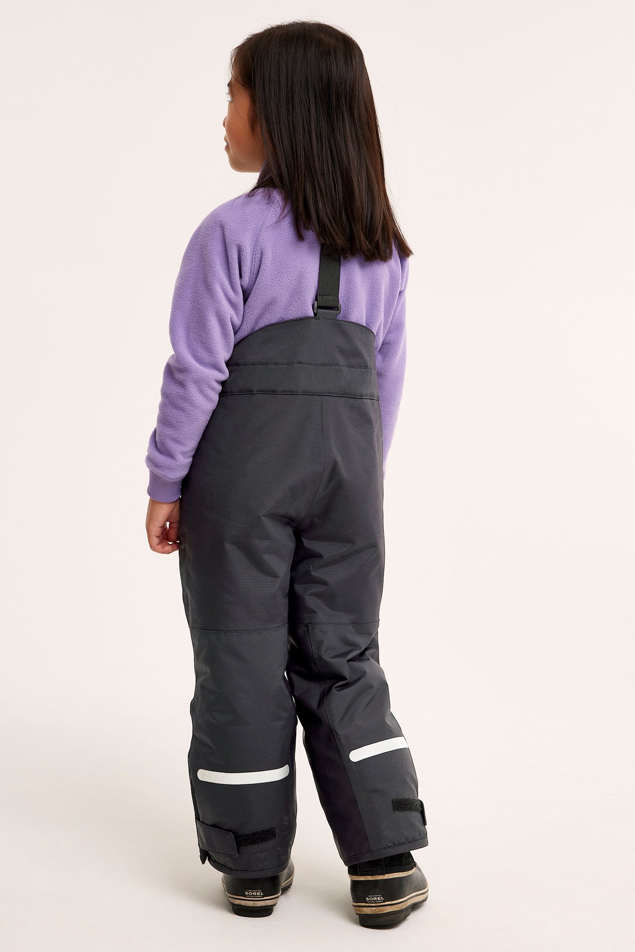Lapsen takakuva mustissa hiihtohousuissa, joissa henkselit, valkoiset heijastinnauhat, violetti fleece ja mustat kengät.