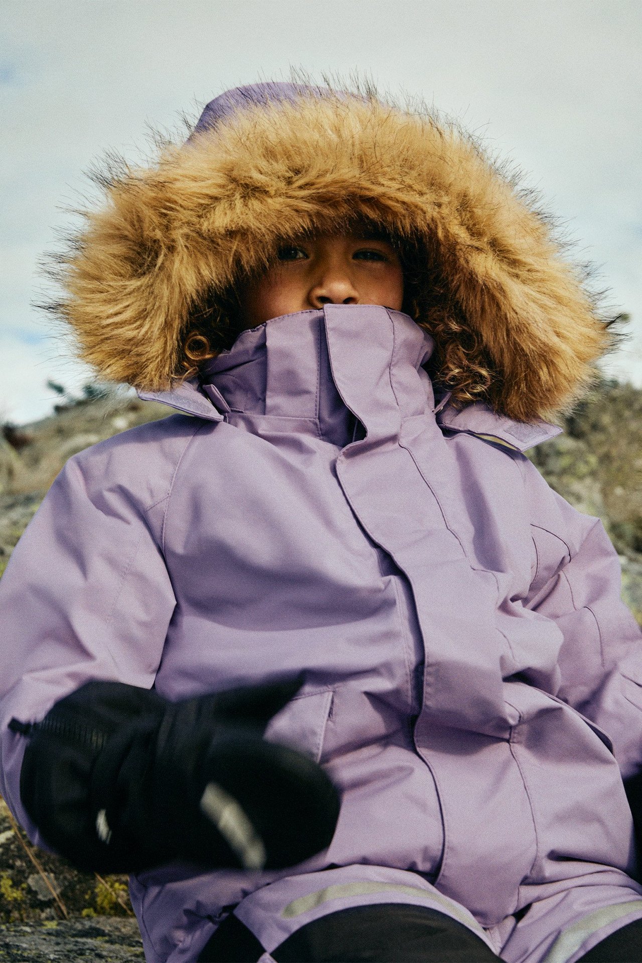 Lähikuva edestä: lapsi yllään lila talvitakki tekoturkisreunaisella hupulla ja mustilla rukkasilla.