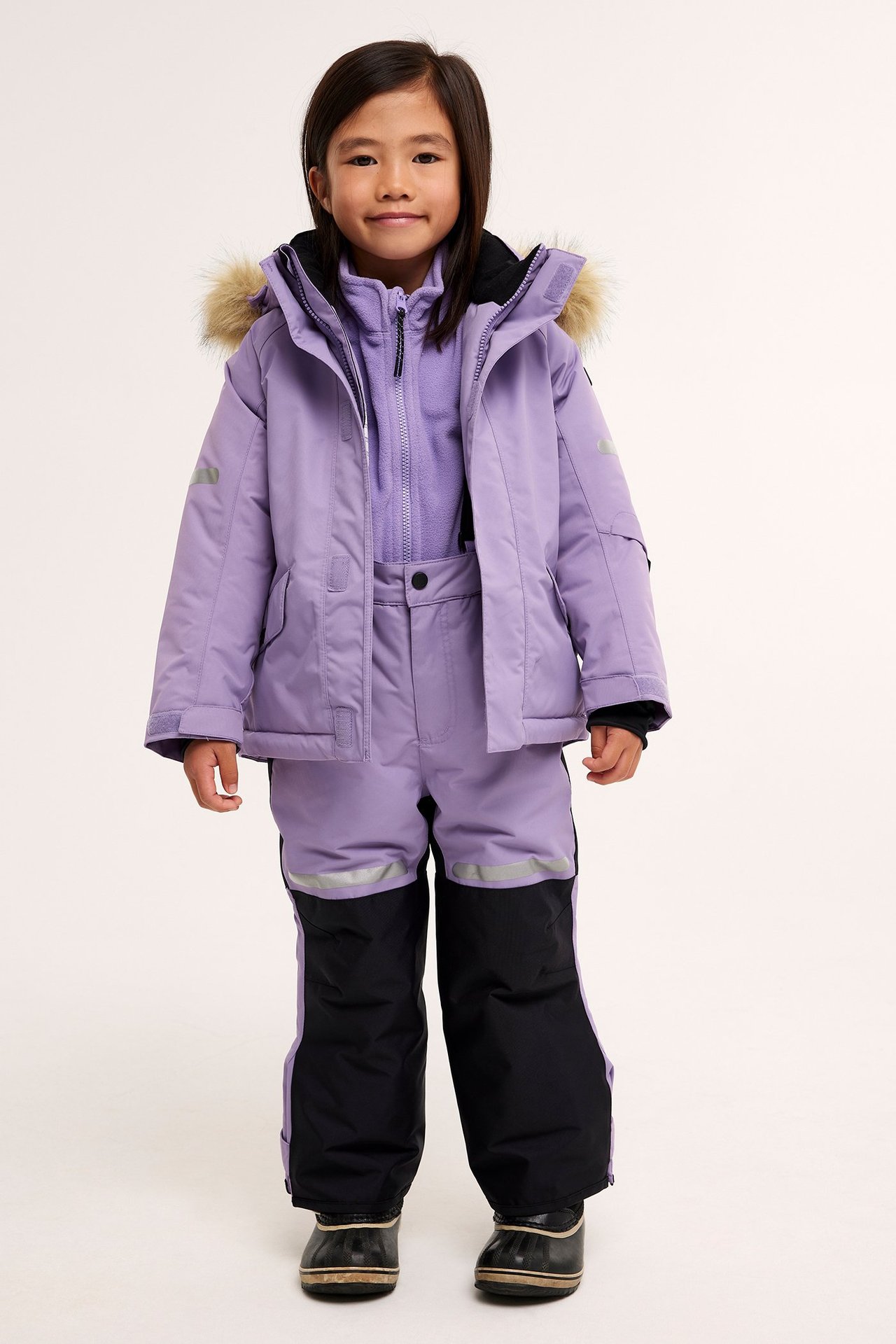 Edestäpäin lapsi lila talvitakissa, jossa turkishuppu, lila fleece ja lila/mustat talvihousut.