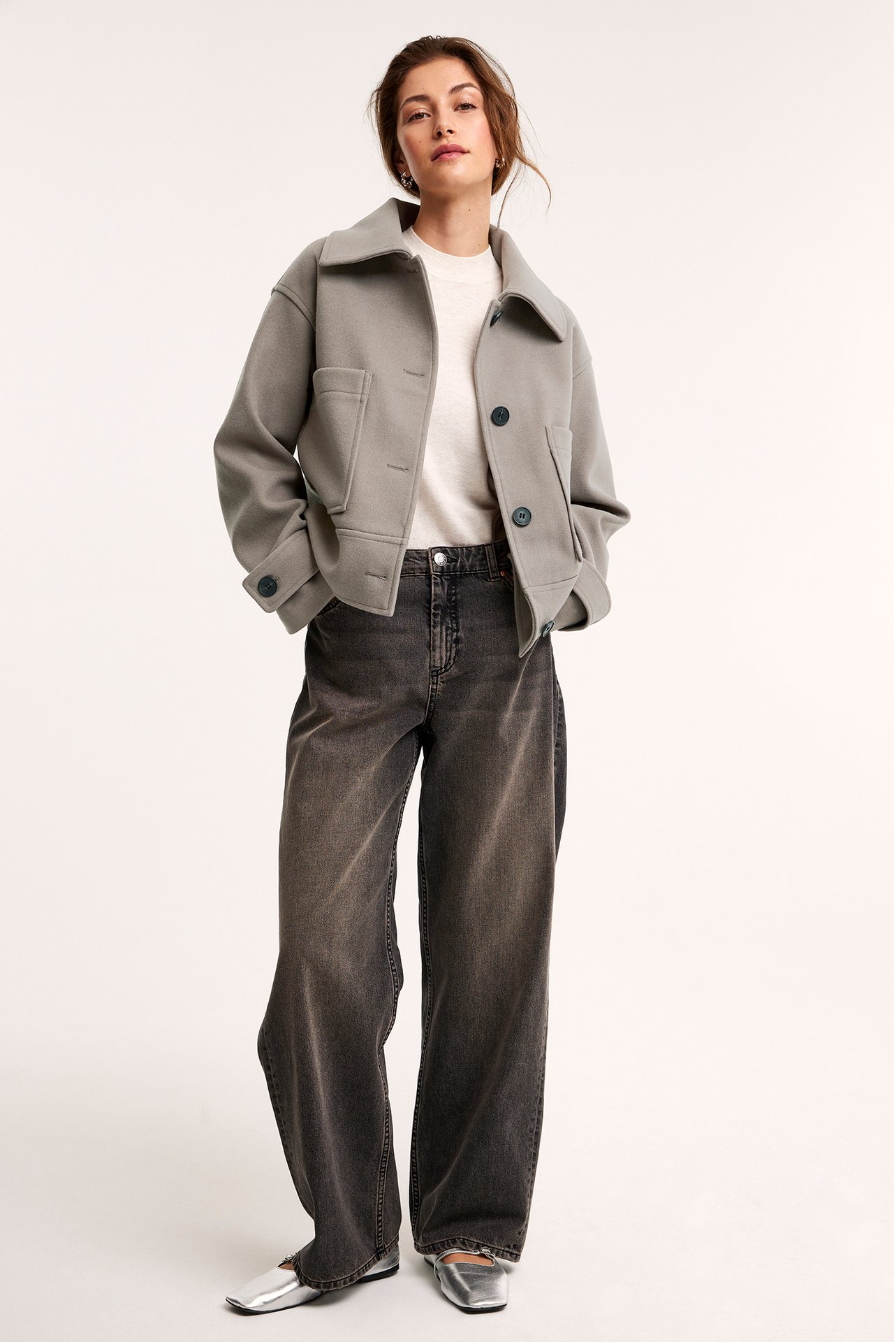 Kvinne har på seg en kort melert beige jakke med krage, lommer, hvit topp, mørkegrå jeans, sølvsko.